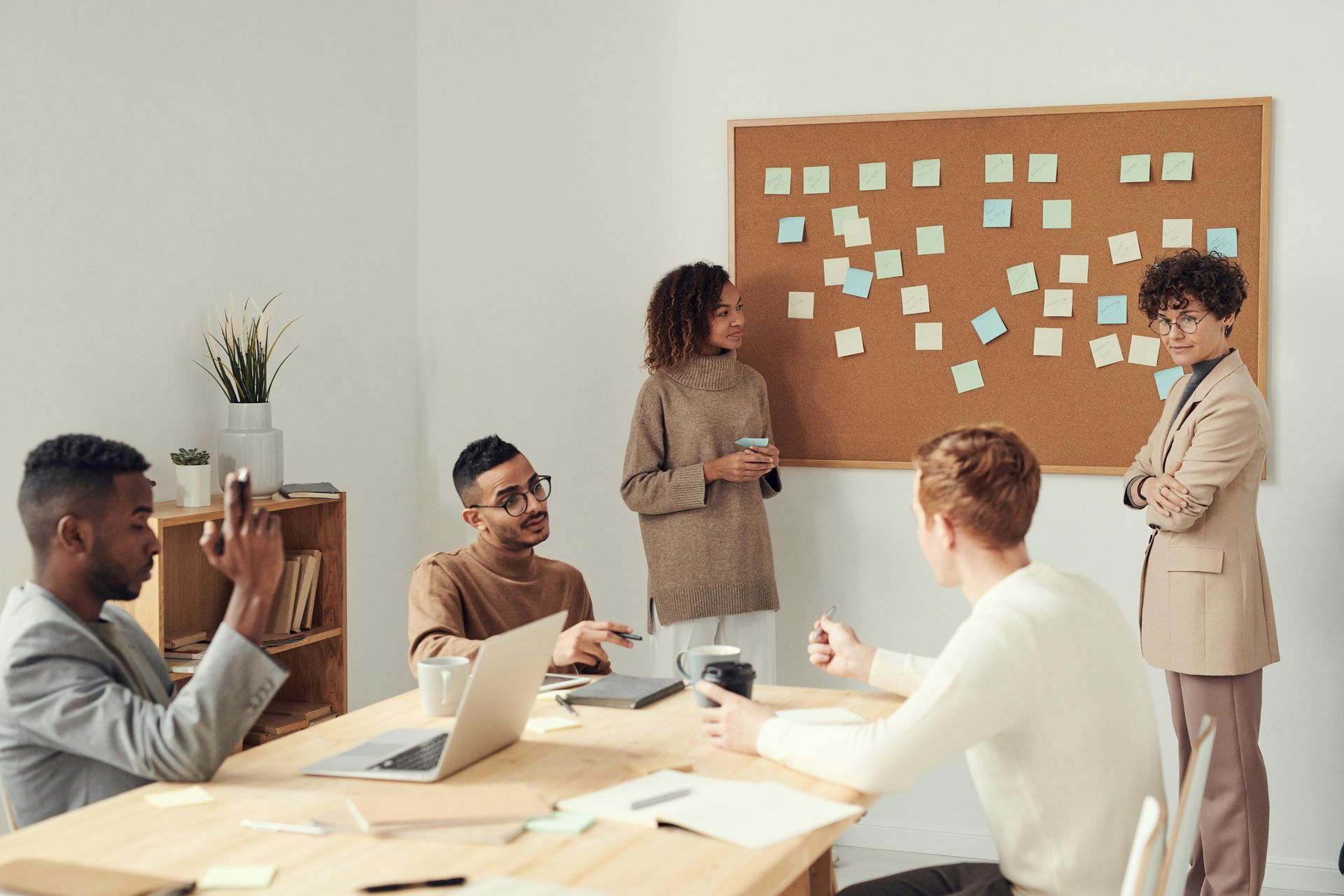Group of people collaborating around a table with sticky notes on a corkboard in a bright office.