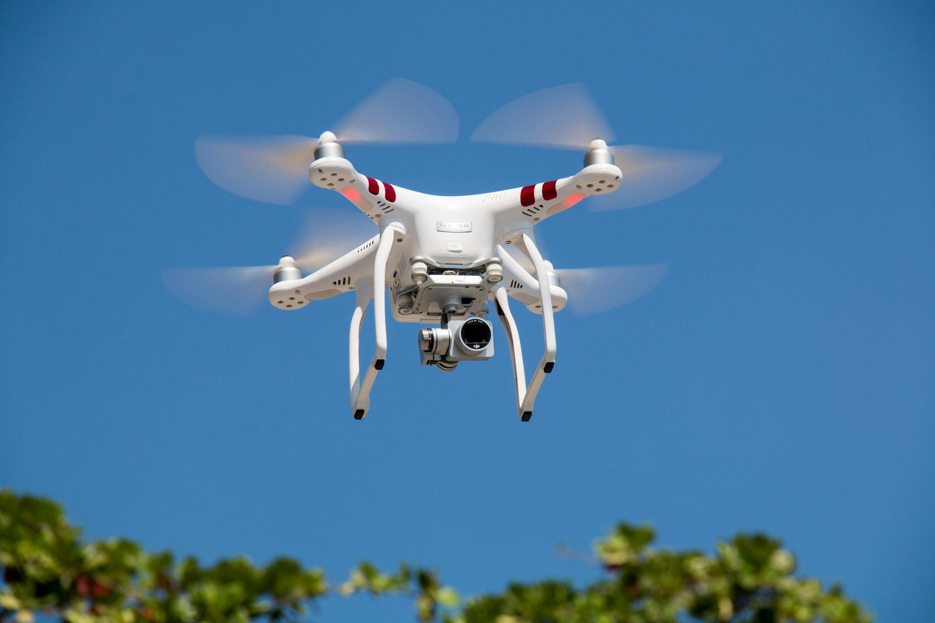 White drone in mid-air against a blue sky, camera attached, blades spinning.