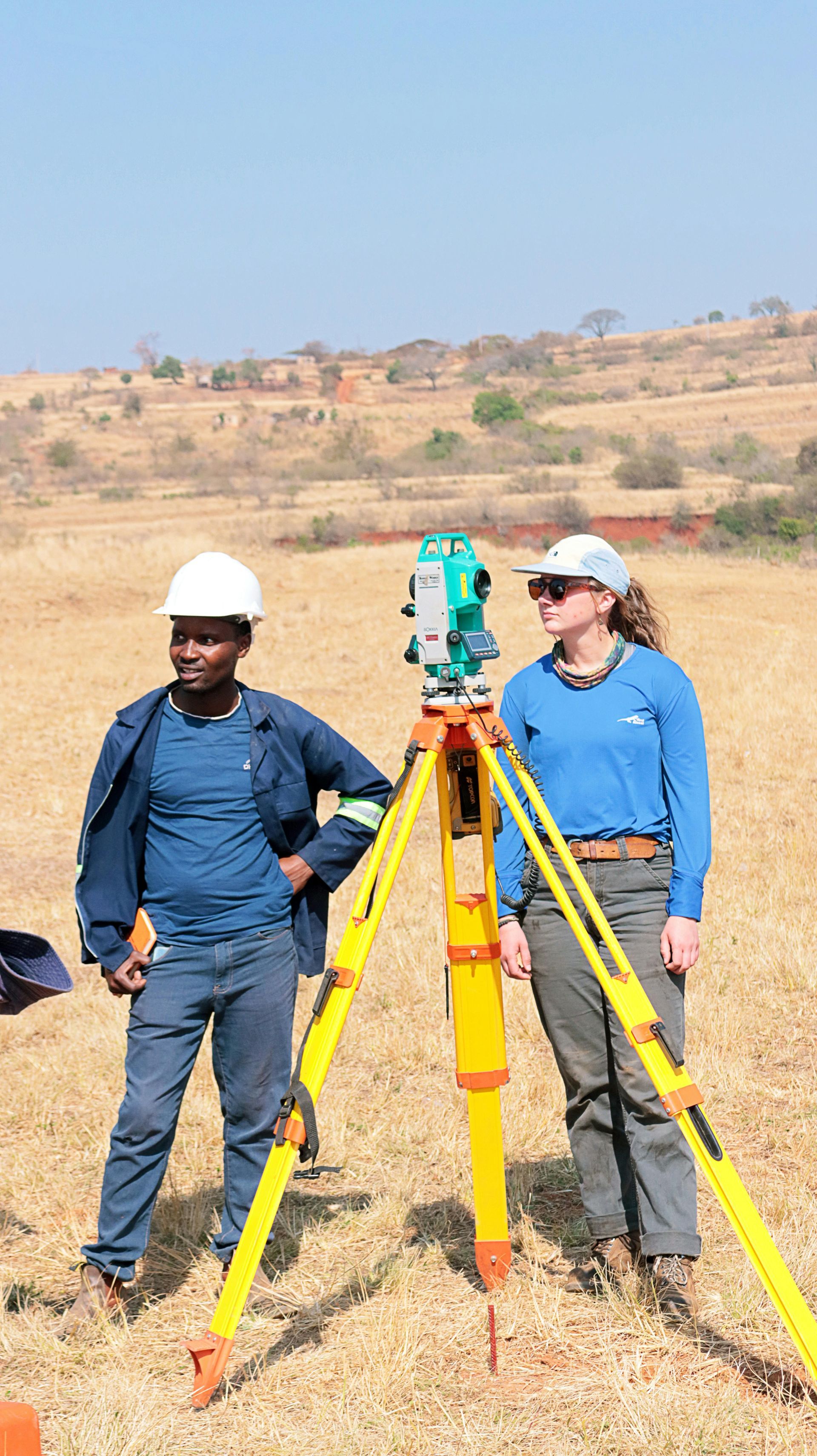 Two people surveying land with a theodolite. One wears a hard hat, the other sunglasses. Outdoor, sunny.