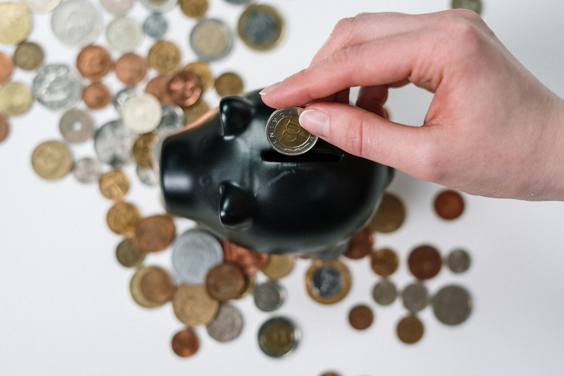 A hand placing a coin into a black piggy bank, surrounded by various coins on a white surface.