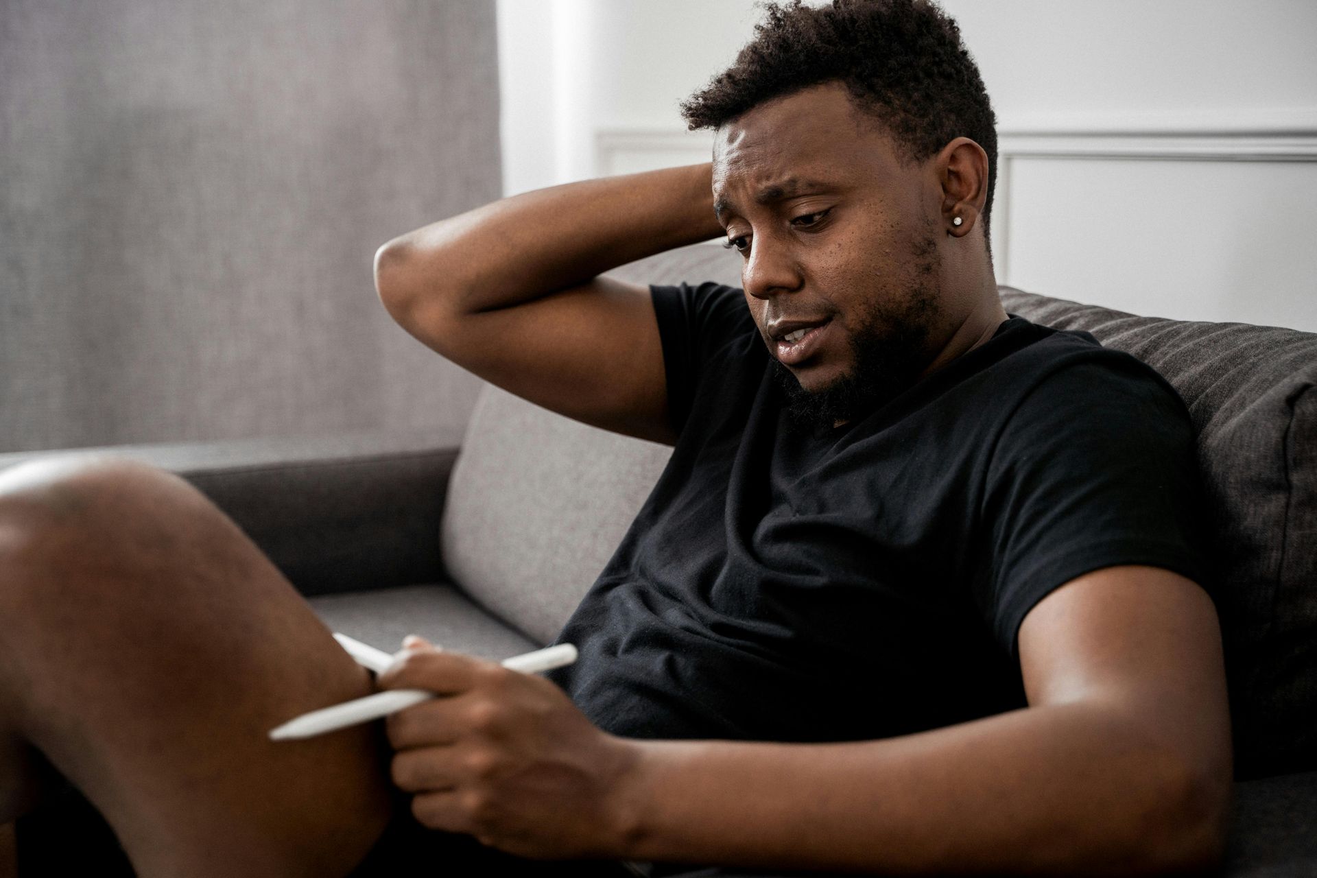 Man with dark skin, wearing a black shirt, sitting on a couch and holding a pen, looking stressed.