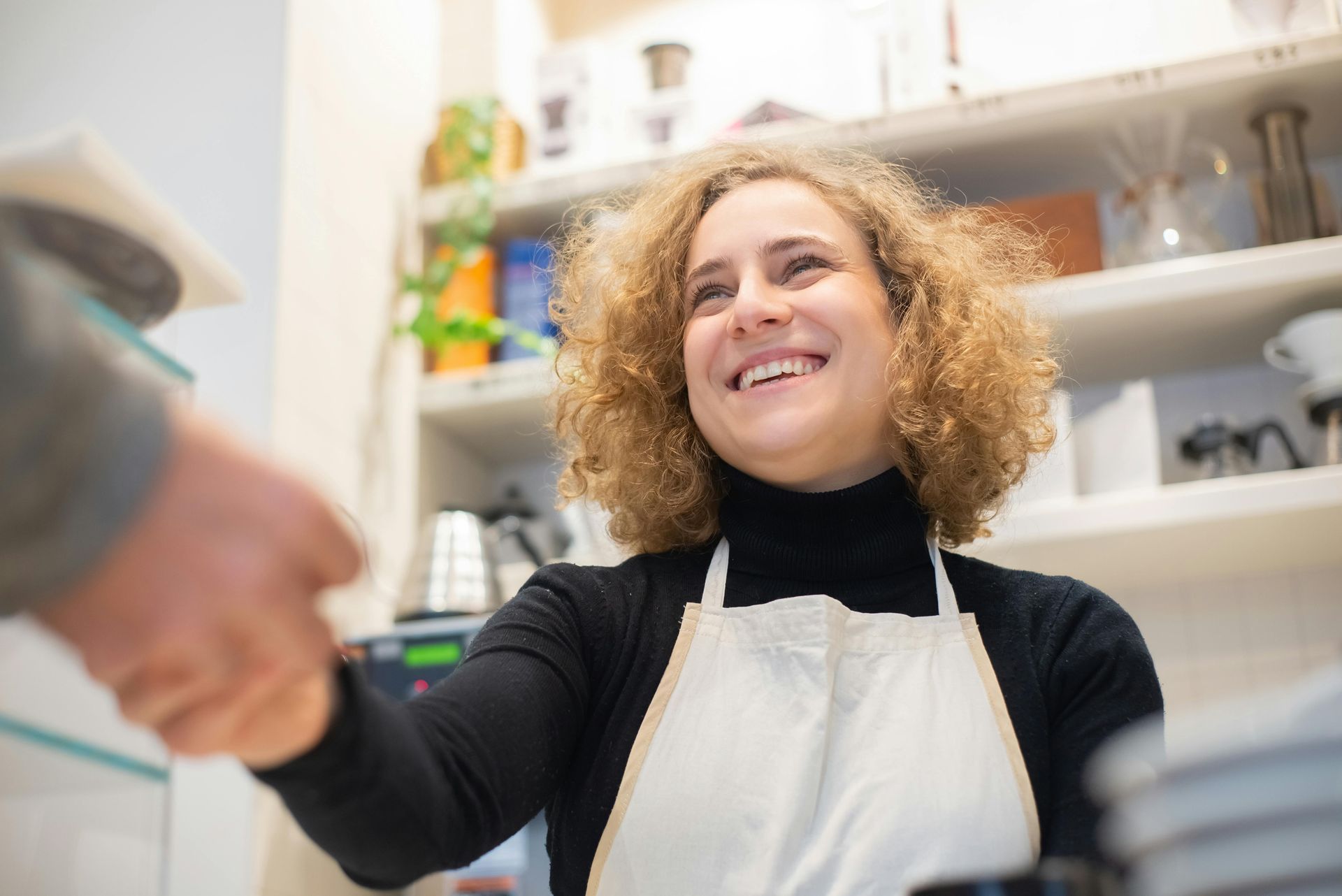 Woman in apron smiles, handing an item to someone, in a cafe setting.