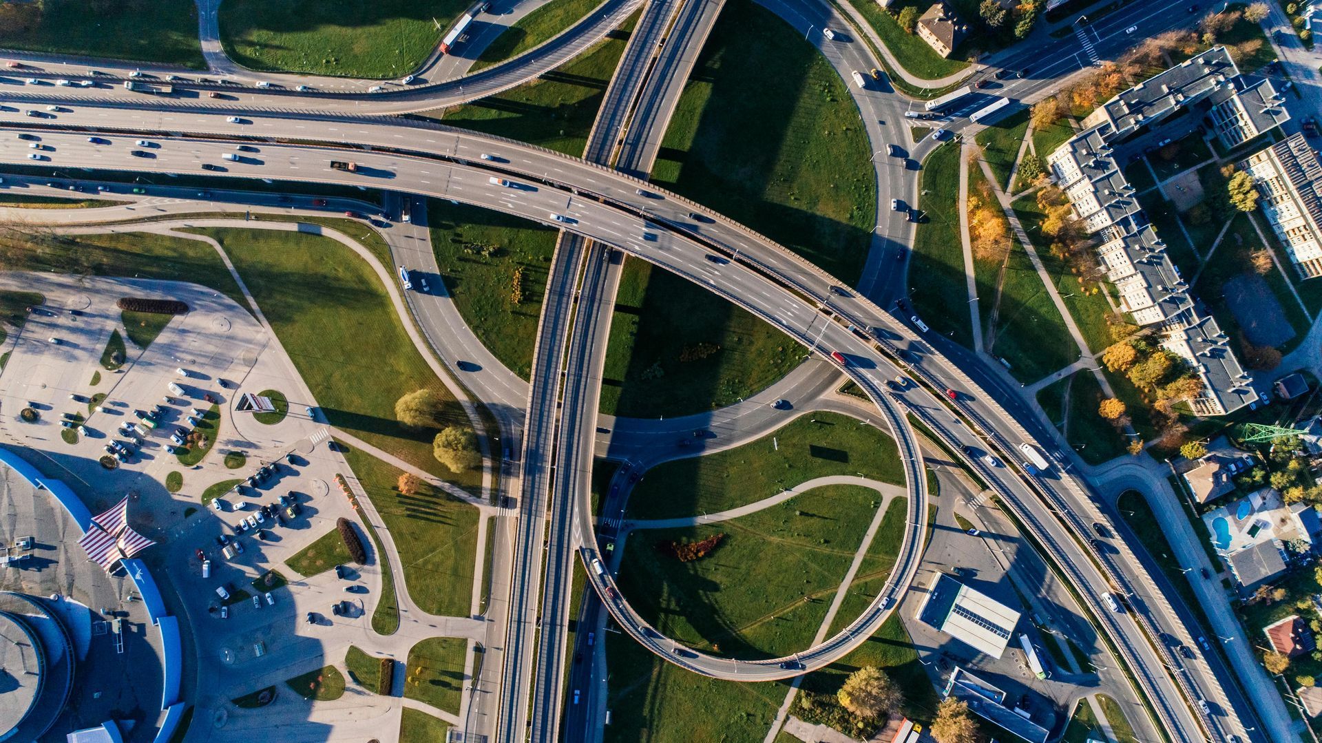 Aerial view of a multi-level highway interchange with cars, green grass, and buildings under a blue sky.