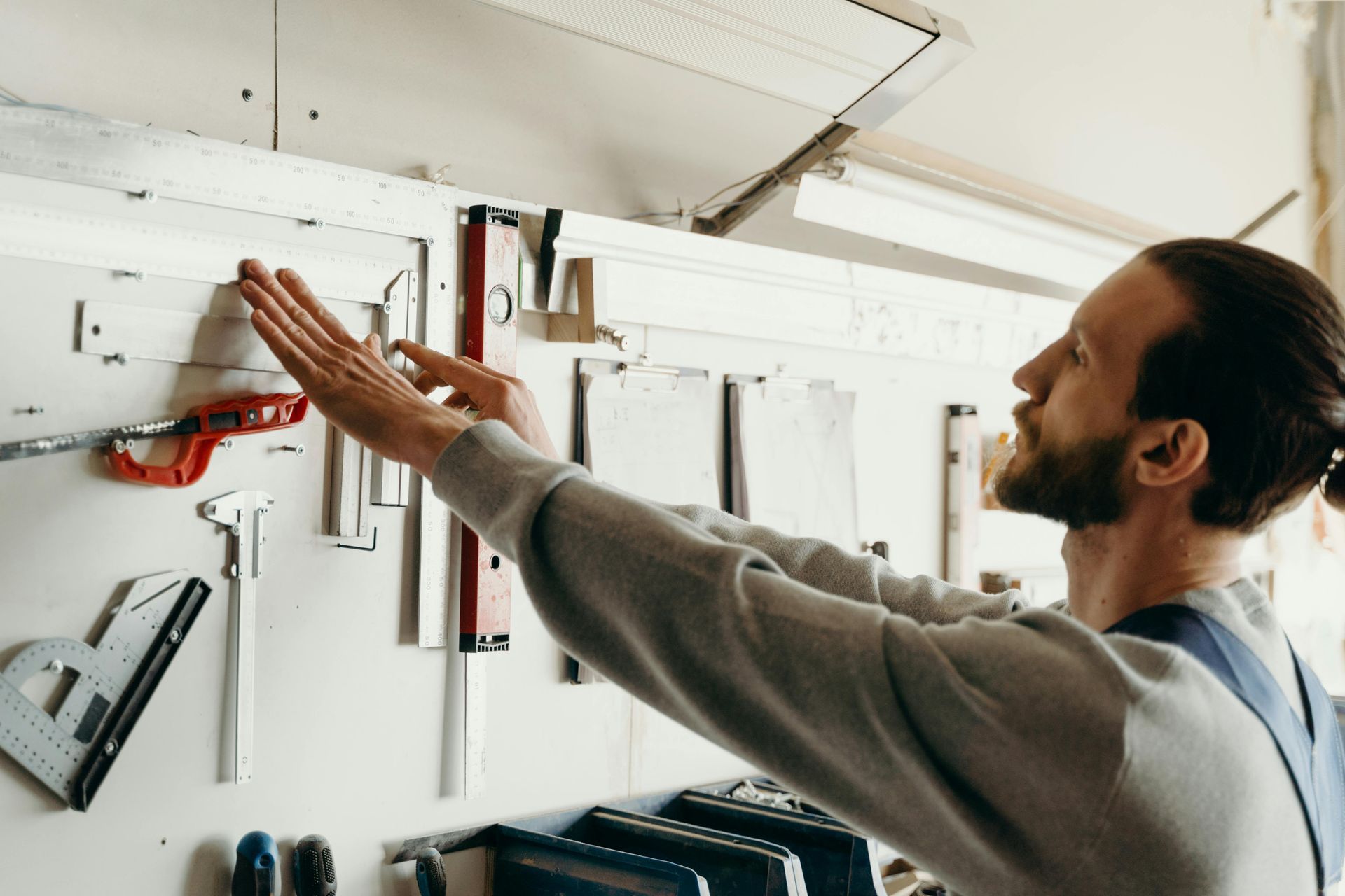 Man in workshop reaching for a tool on a pegboard, inspecting tools.