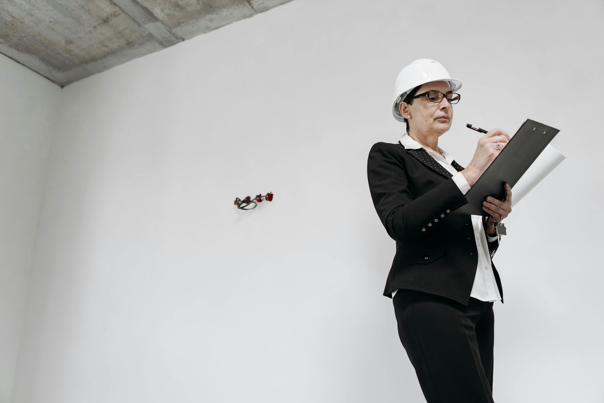 Woman in hard hat, suit, and glasses, inspecting a construction site, taking notes on a clipboard.