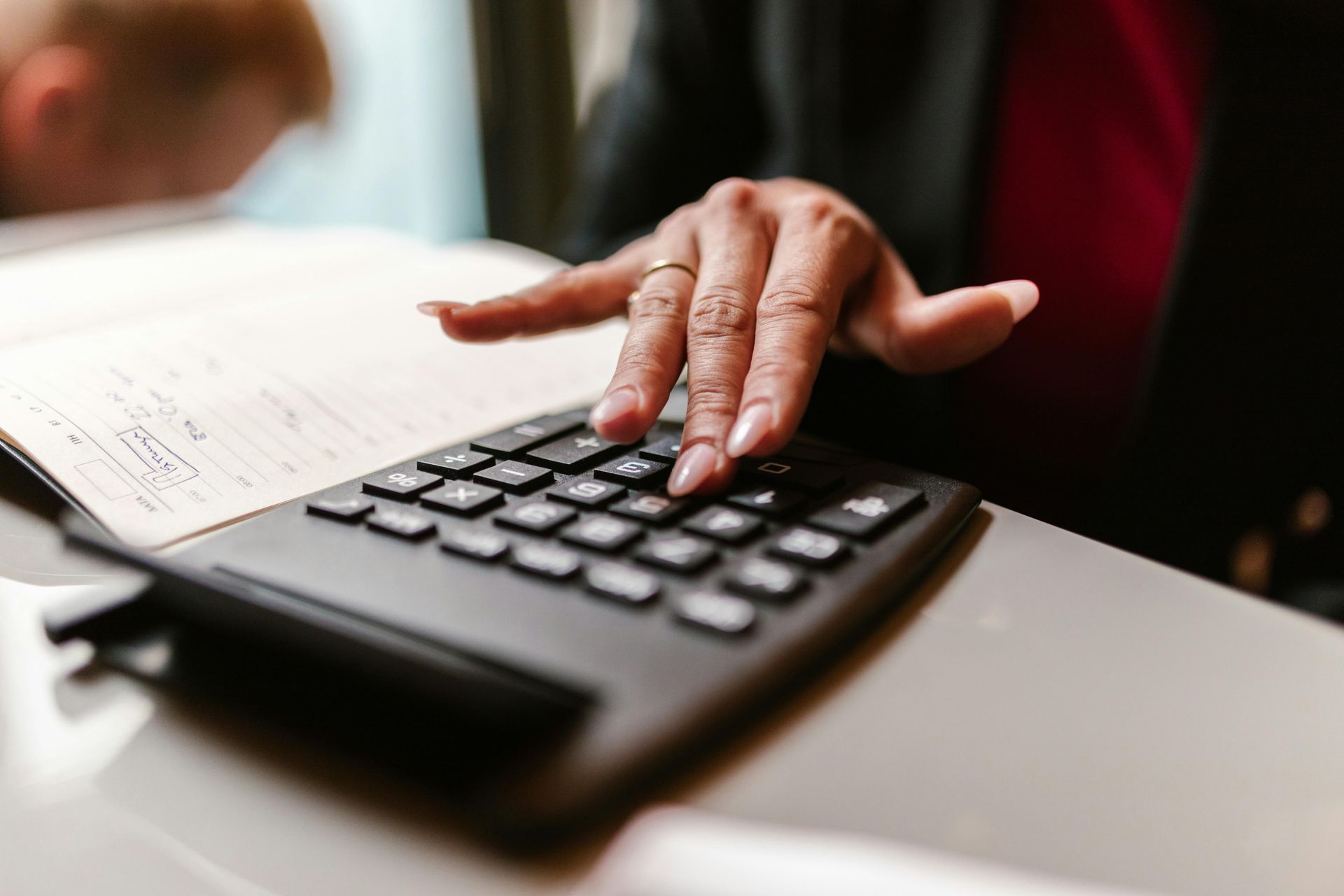 Woman's hands on a black calculator, calculating numbers next to an open book, at a desk.