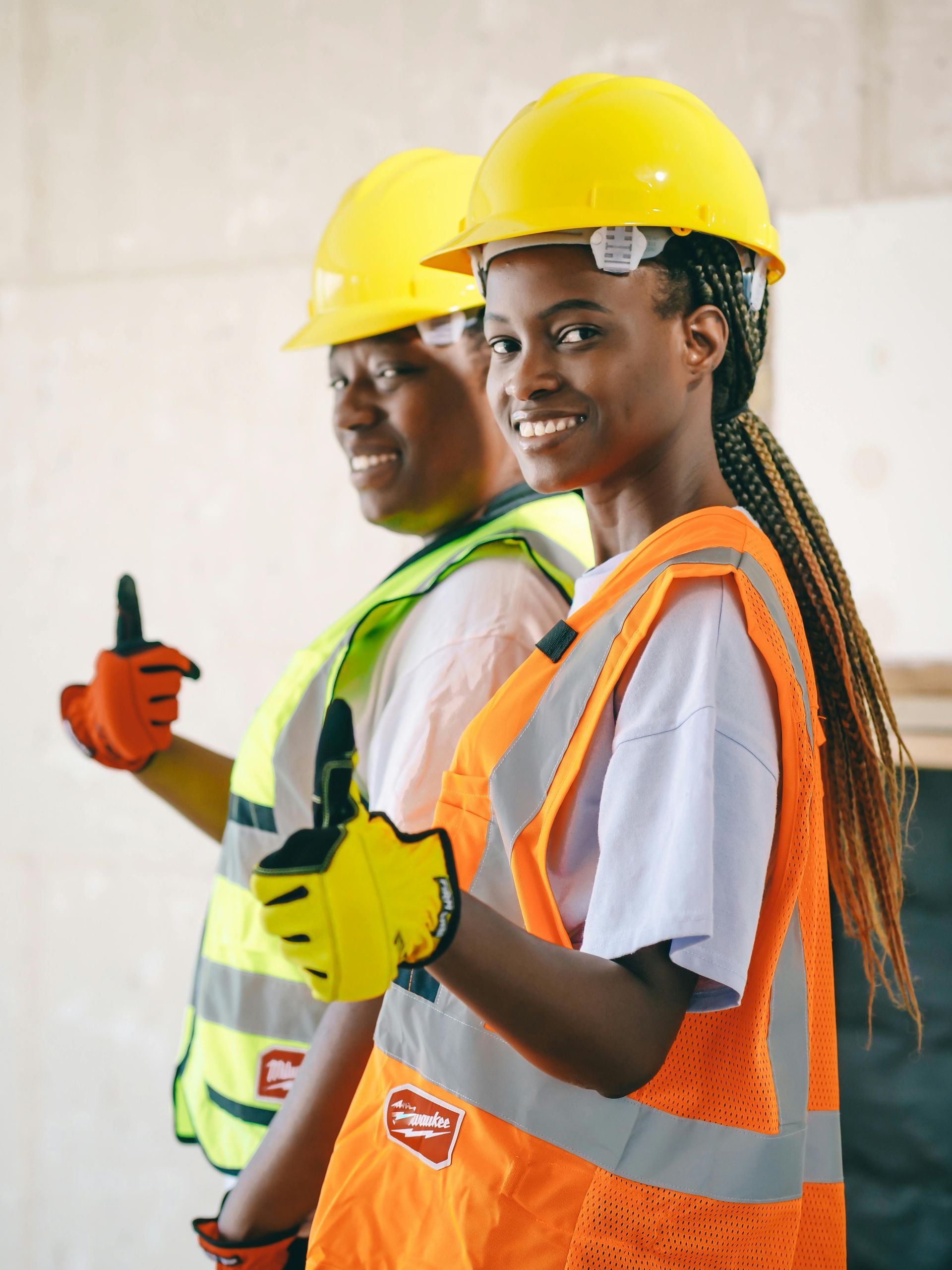 Two construction workers, wearing hard hats and vests, give a thumbs-up.