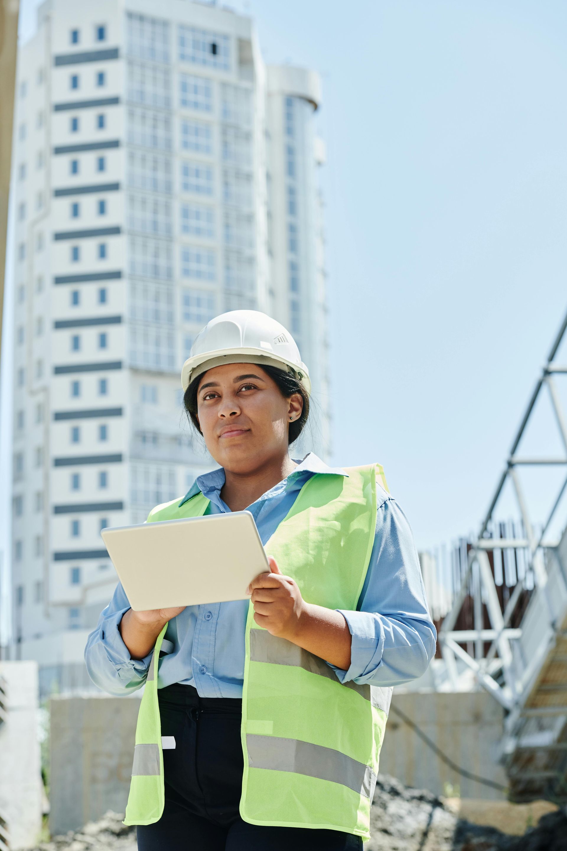 Woman in hard hat and vest, holding tablet on a construction site, in front of a high-rise building.