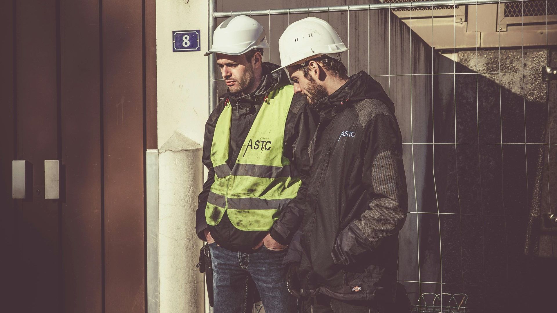 Two construction workers wearing hard hats and safety vests are standing next to each other.