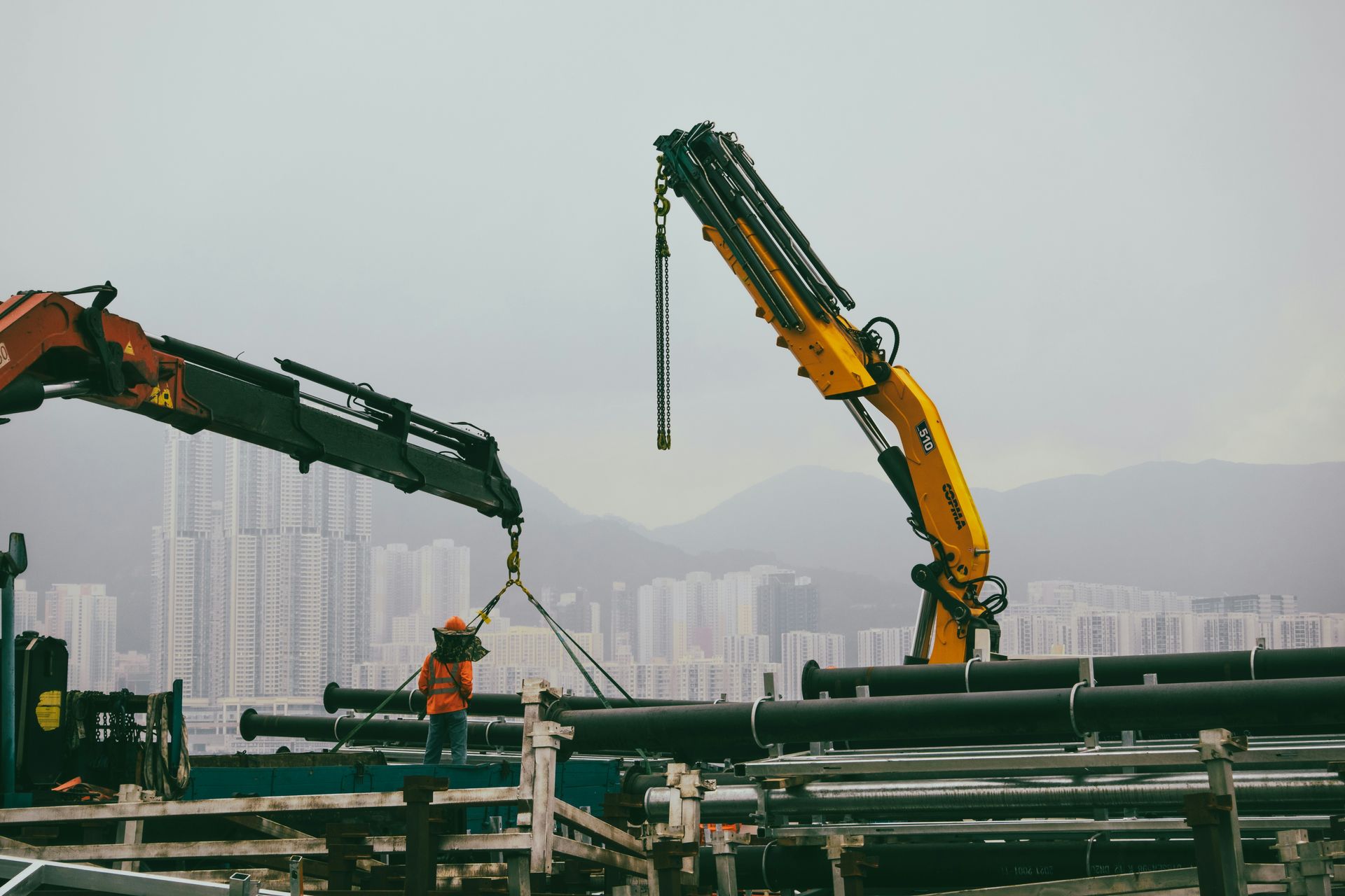 Construction site: worker guides crane lowering pipes. Yellow and red cranes, city skyline backdrop.