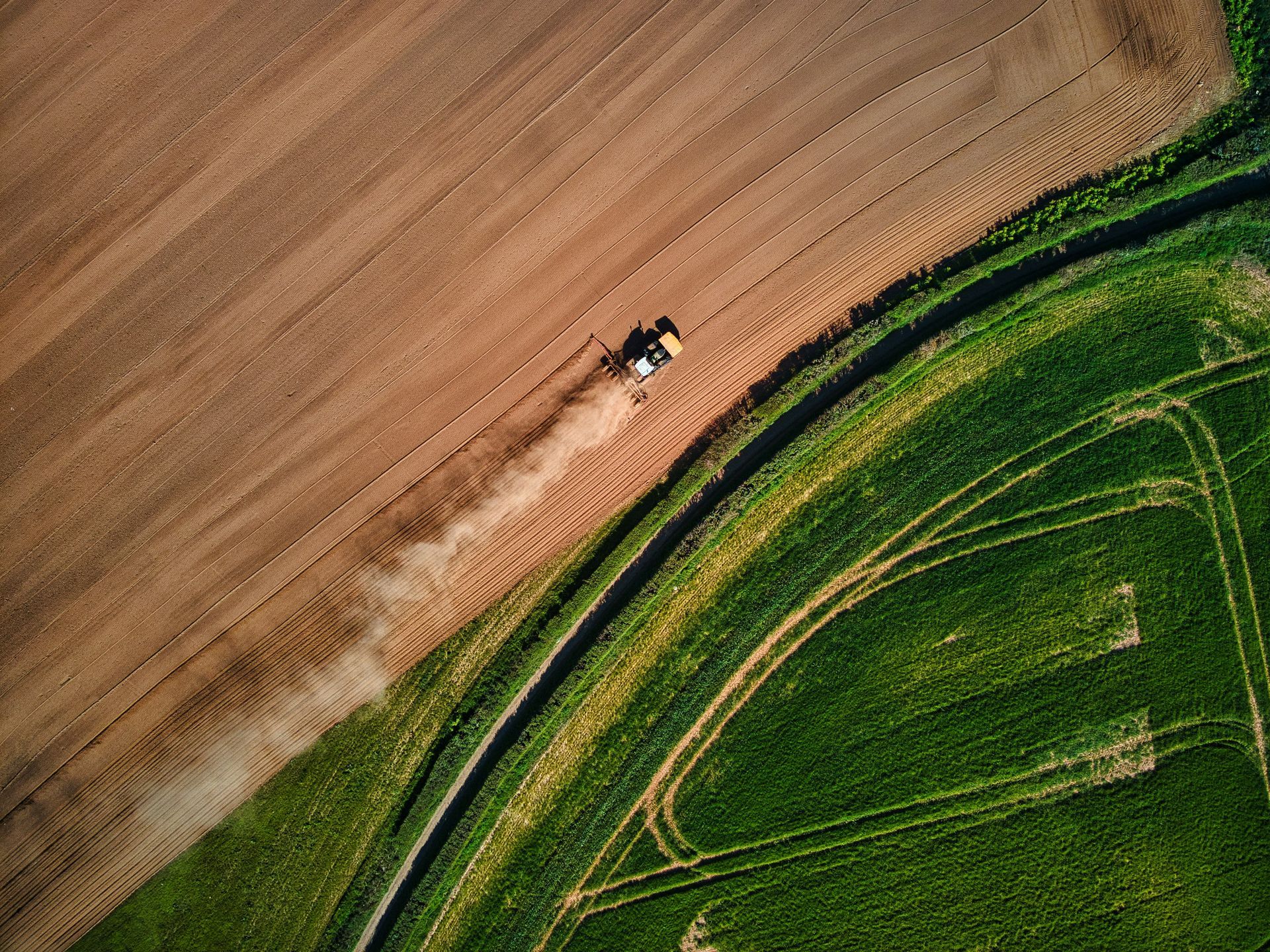 Tractor plowing a brown field, next to a green field with a winding path.