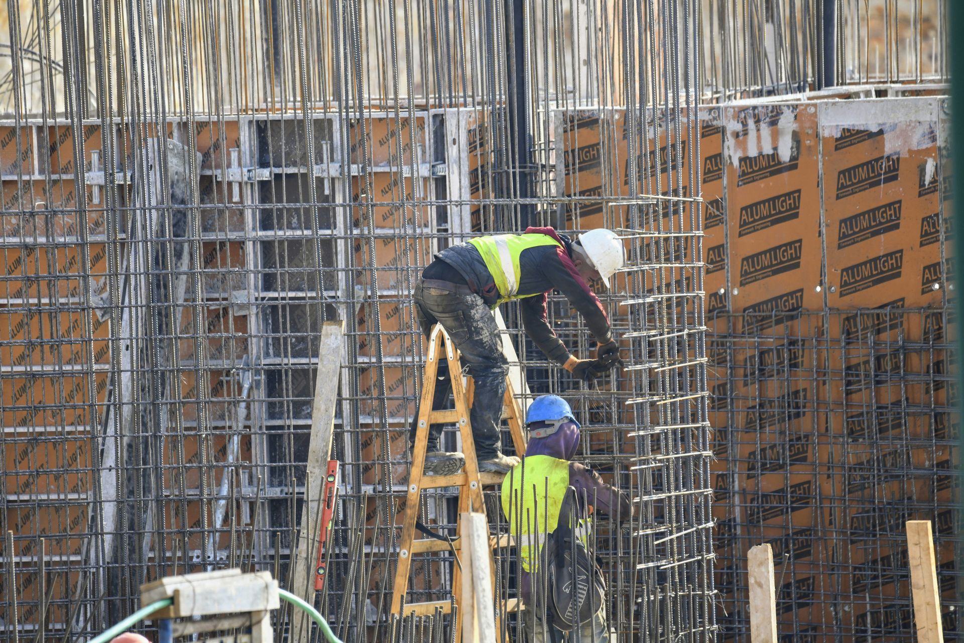 Construction workers on a ladder, working with rebar at a building site; concrete forms in background.