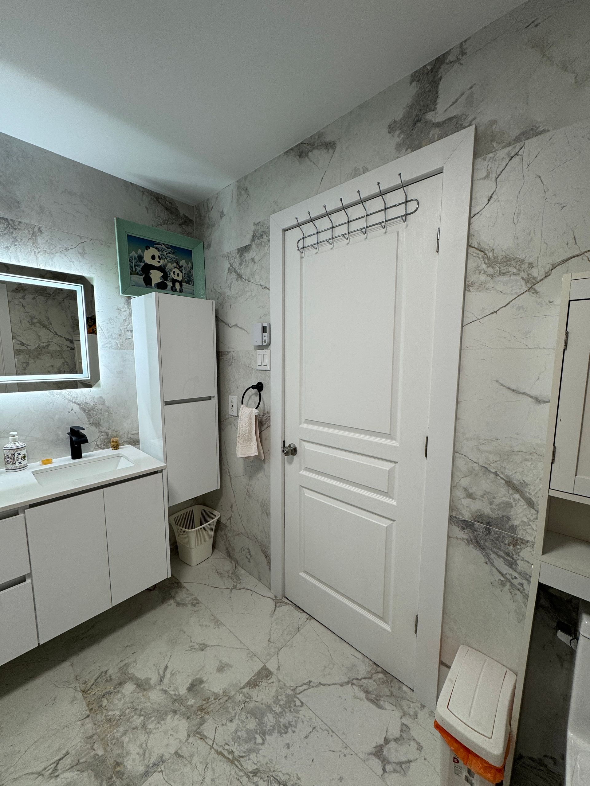 Une salle de bain avec des murs en marbre, des armoires blanches, un lavabo, un miroir et une porte.