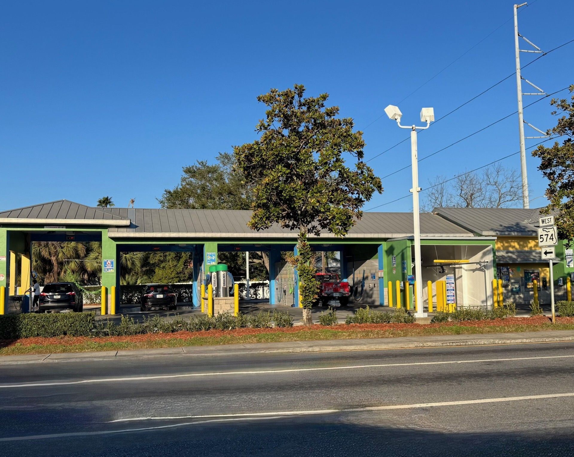 A car wash with multiple bays, a light-colored building with green, yellow and blue accents, a blue sky, and a large tree.