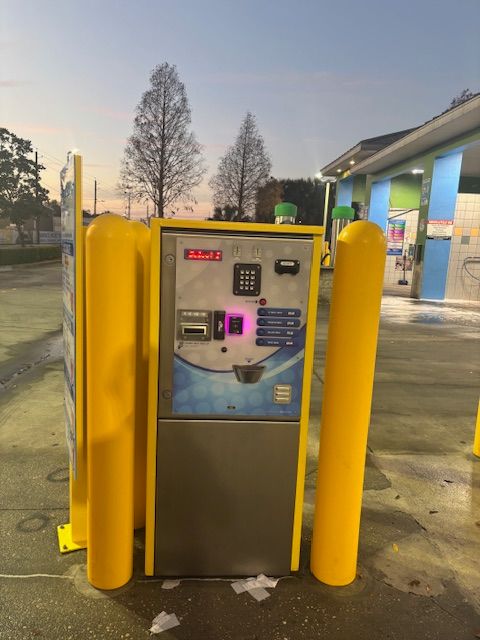 Car wash payment kiosk with yellow bollards. Silver and gray machine with a touch screen. Evening setting.