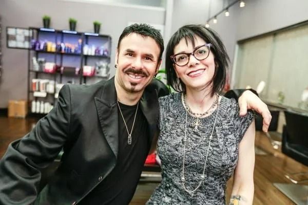 Two people smiling and posing indoors at a bar or lounge, with shelves of bottles in the background.