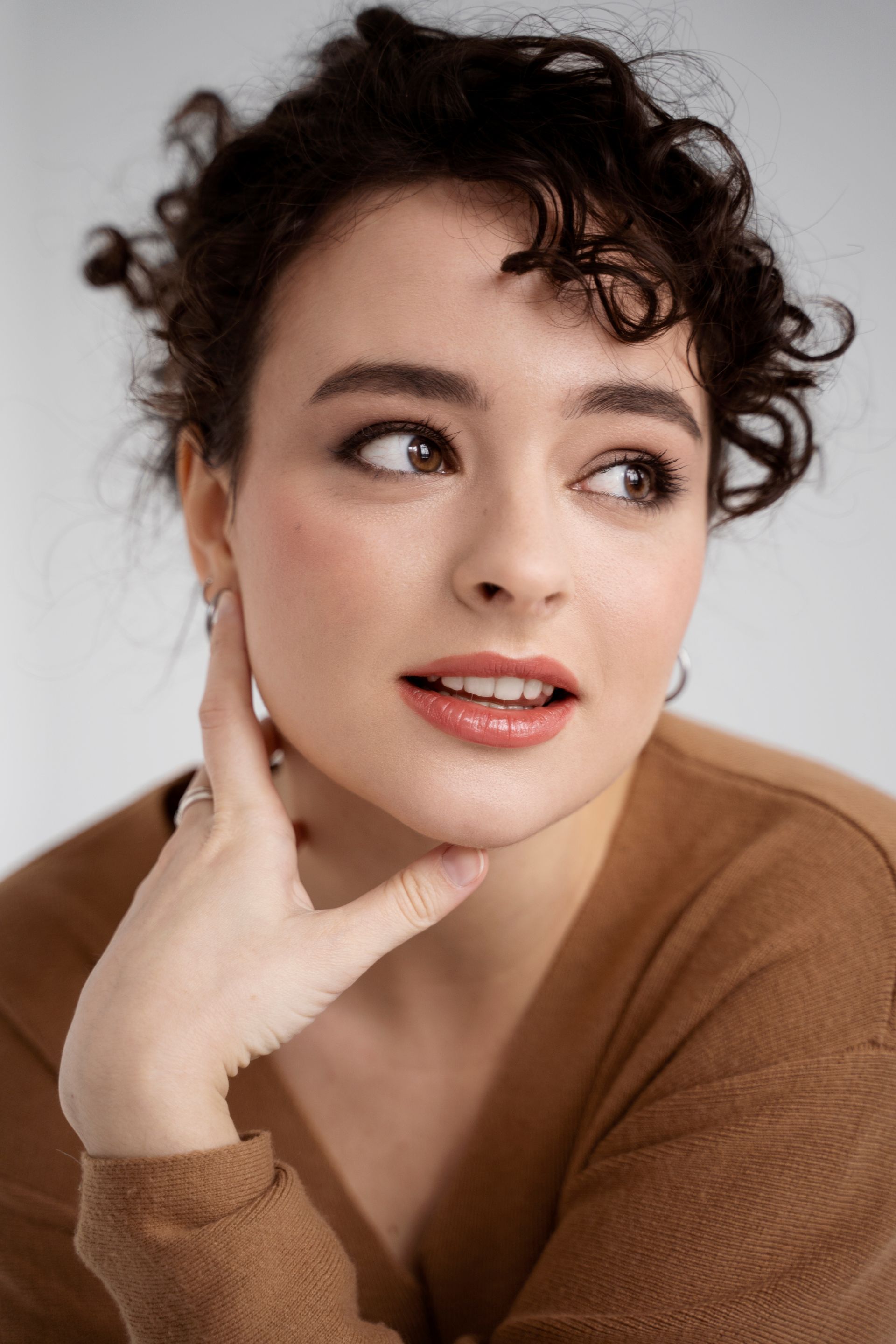 A woman with curly hair is wearing a brown sweater and earrings.
