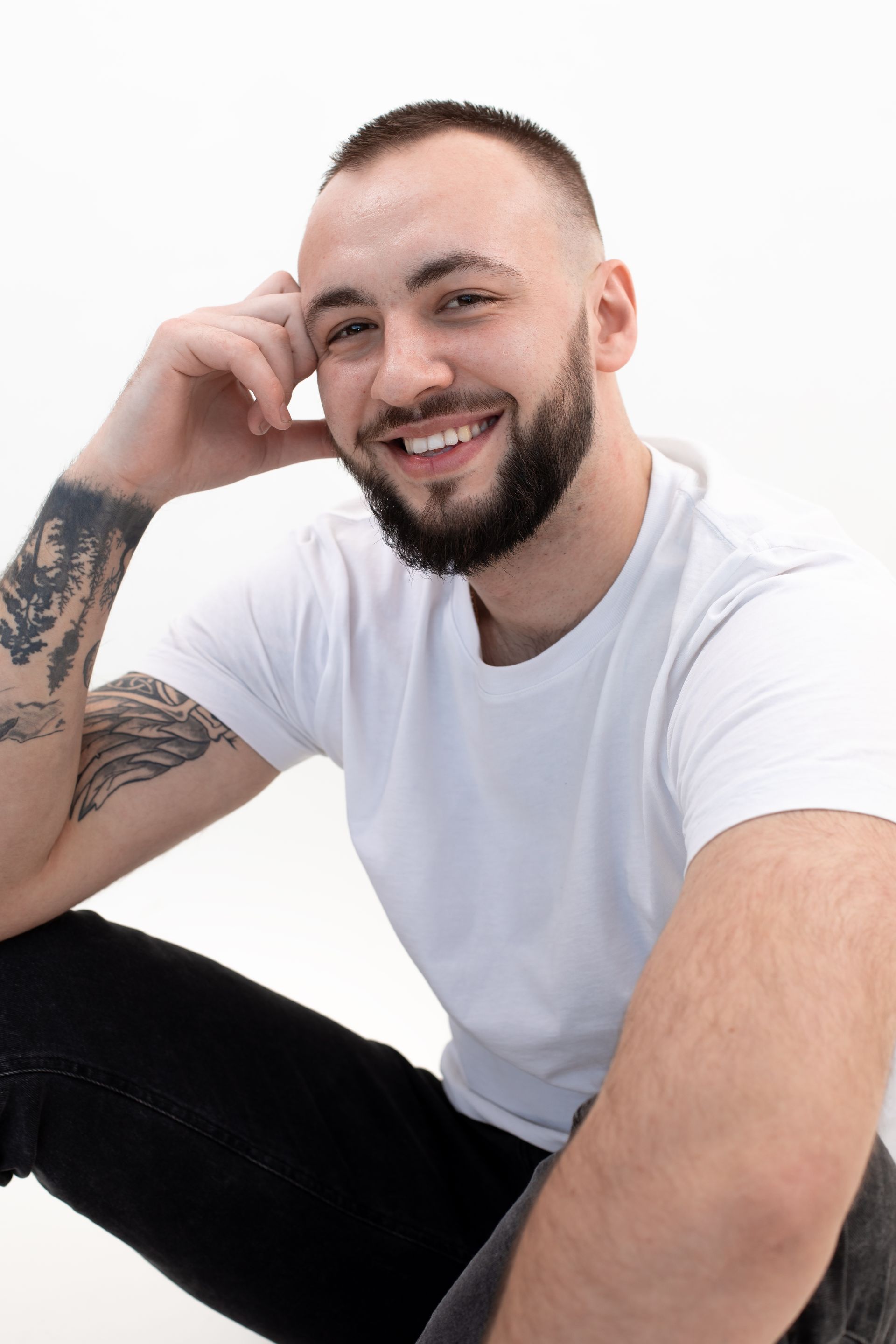 A man with a beard and tattoos is sitting on the floor and smiling.