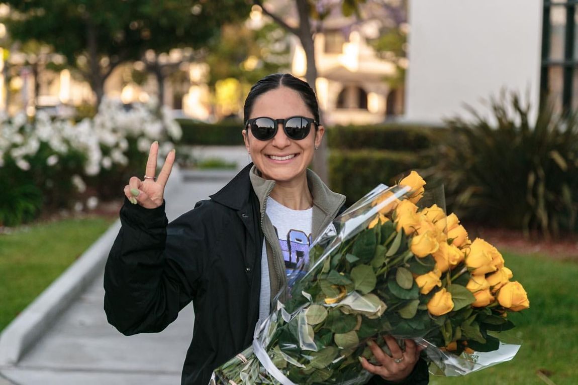 A woman is holding a bouquet of yellow roses and giving a peace sign.
