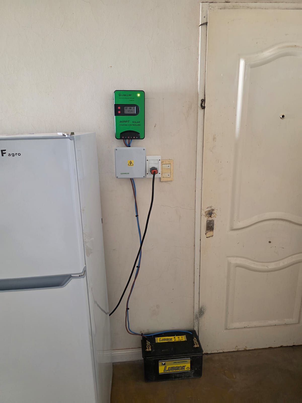 Sistema de energía solar instalado en interiores: controlador verde, caja blanca, batería, refrigerador, puerta.