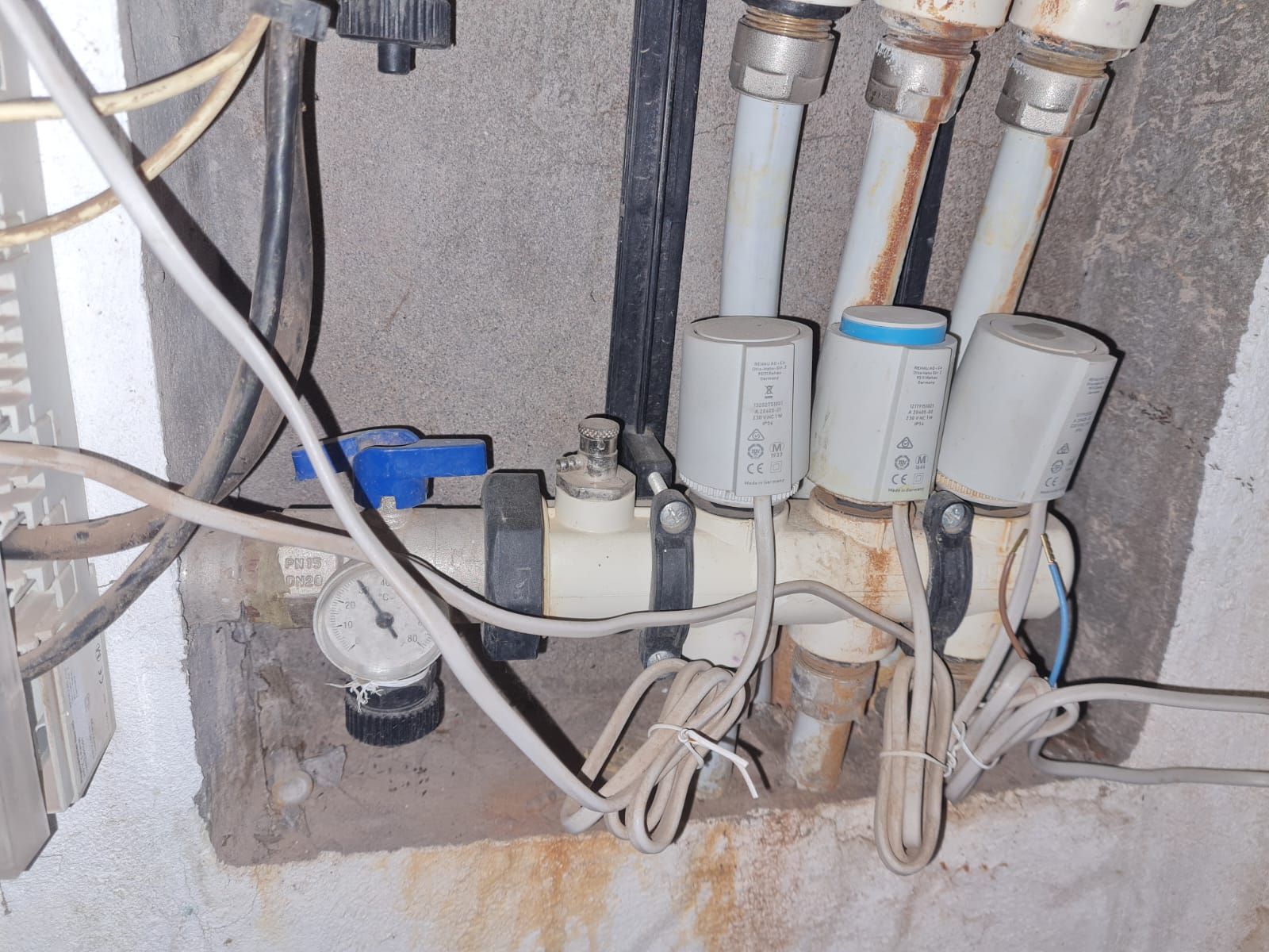 Primer plano de tuberías de plomería con actuadores blancos, una válvula azul y un manómetro montado en una pared.