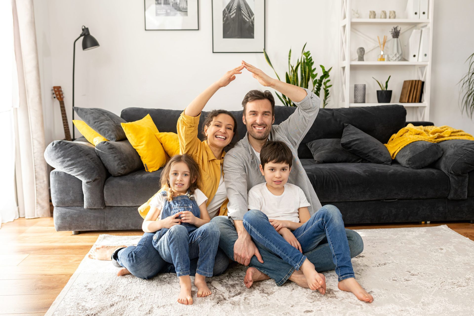 A family sits on a rug in a living room, making a roof shape with their arms above their heads.