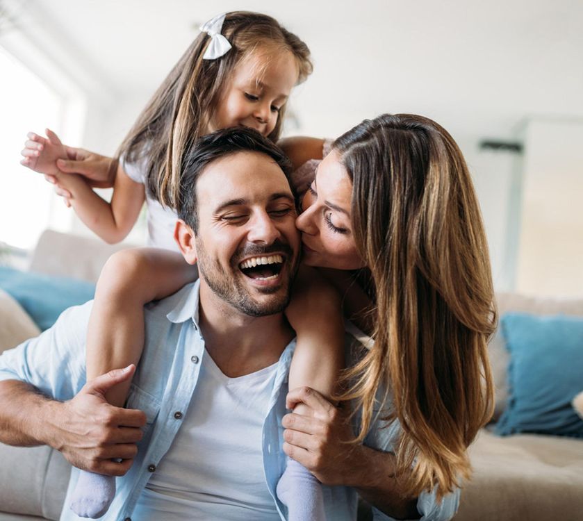 A smiling man carries a child on his shoulders while a woman kisses his cheek indoors.