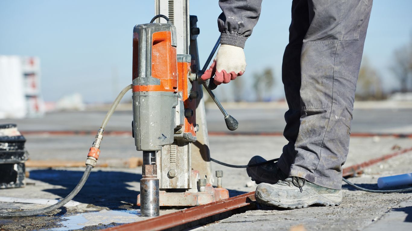 Person in gray work clothes using an orange drill on concrete outdoors.