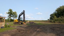 Yellow excavator on a dirt field under a blue sky near trees and a small building.