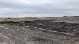 Bare muddy field under a cloudy sky with a line of low shrubs on the horizon