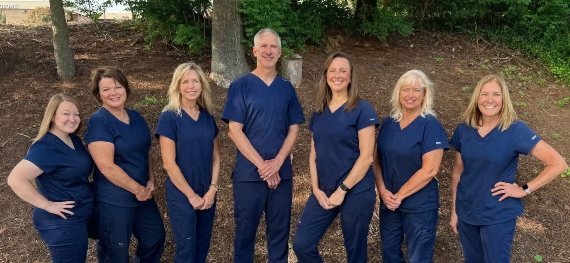 Seven people in matching navy outfits standing outdoors, smiling in front of greenery and stone steps