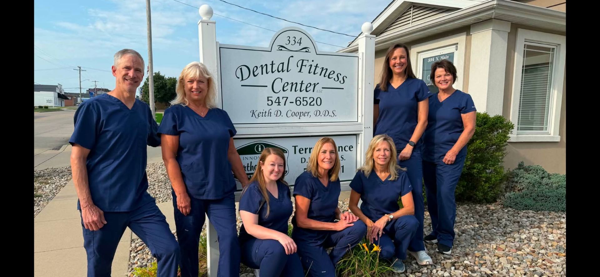 Group of six people posing outside a dental office sign in front of a white building.