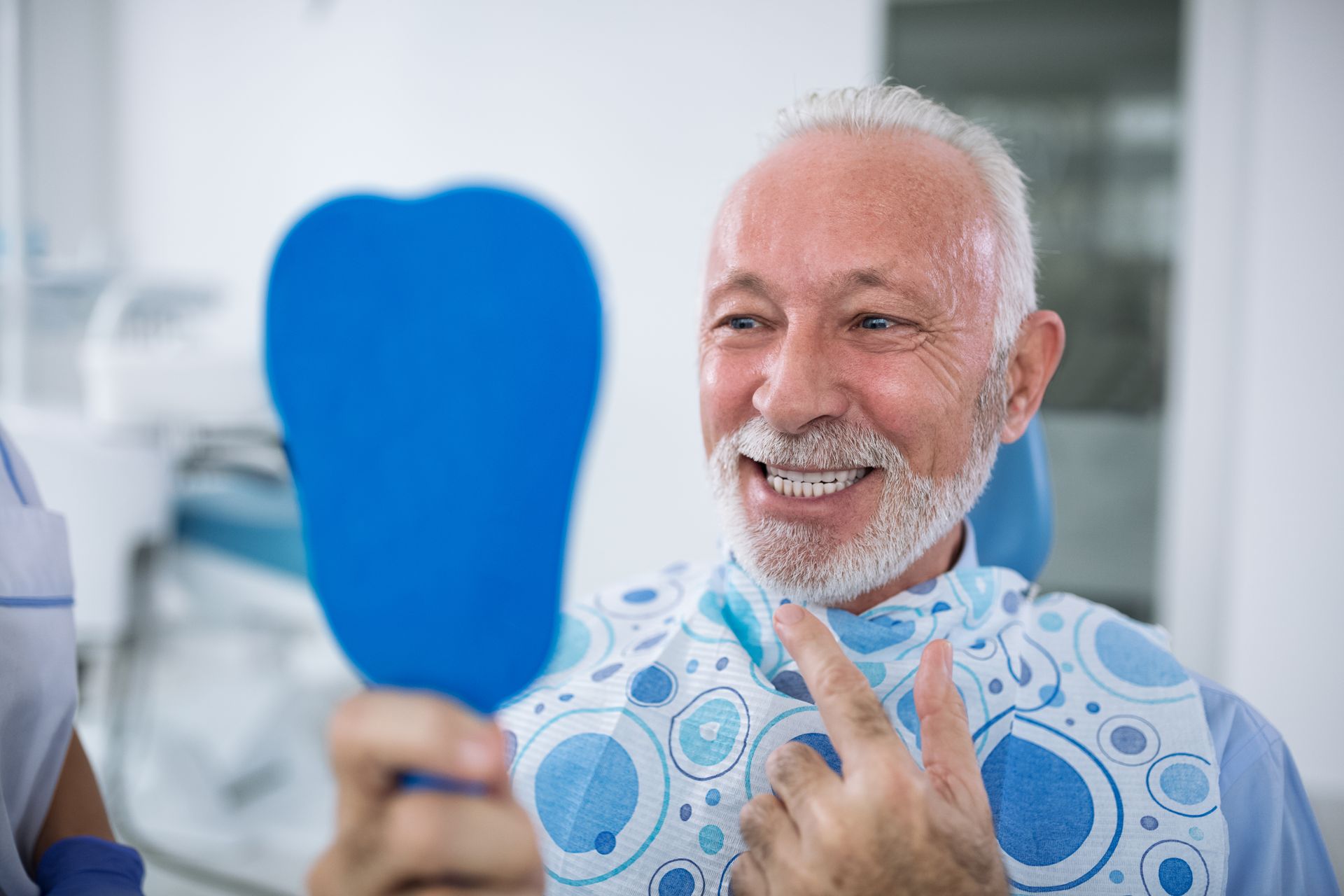 Smiling man in a blue dental bib holding a mirror in a bright clinic.