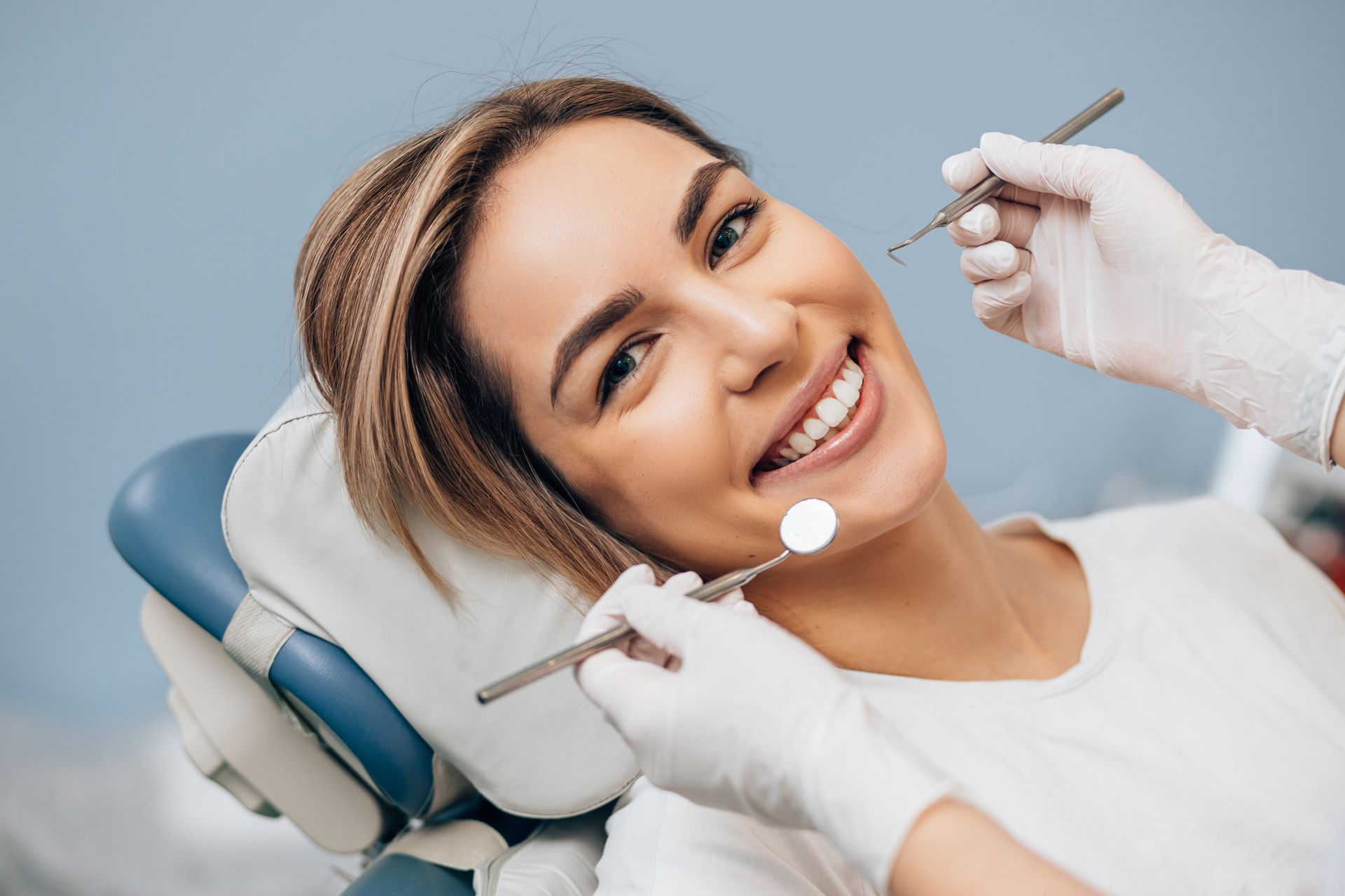 Patient smiling during dental cleaning while dentist uses mirror and tools in a bright clinic.