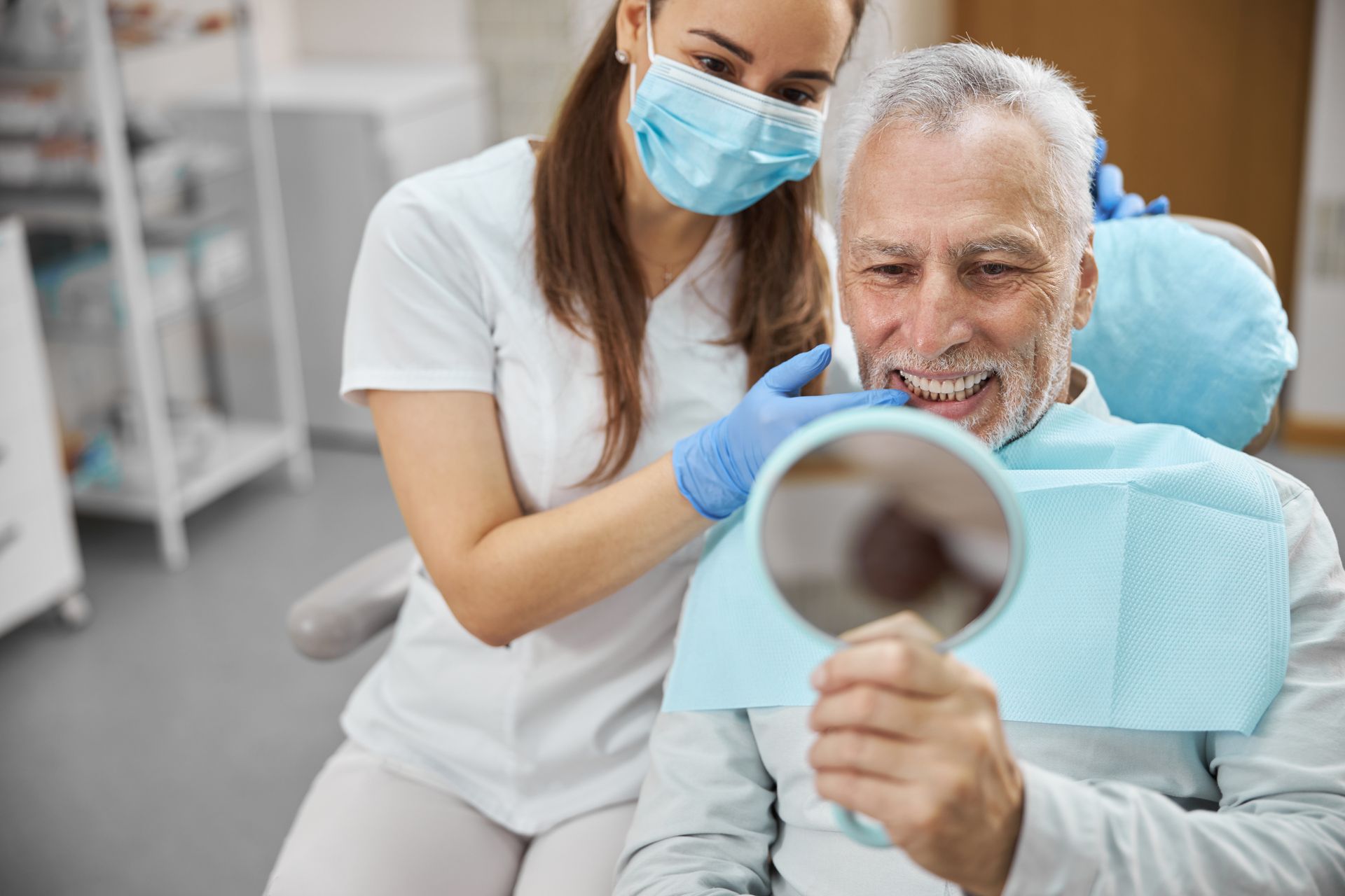 Dentist wearing blue gloves examines a smiling patient with a handheld mirror in a bright clinic.