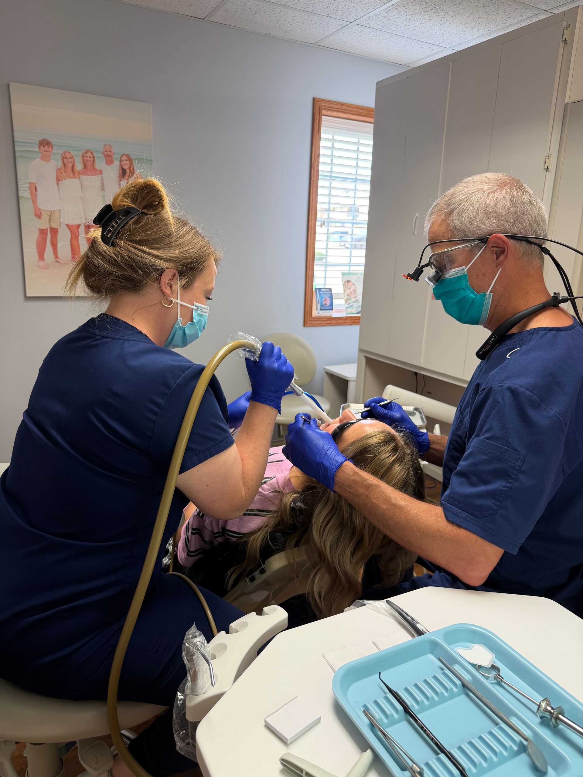 Two dental clinicians in blue scrubs treat a patient in a dental chair, with tools on a nearby tray.