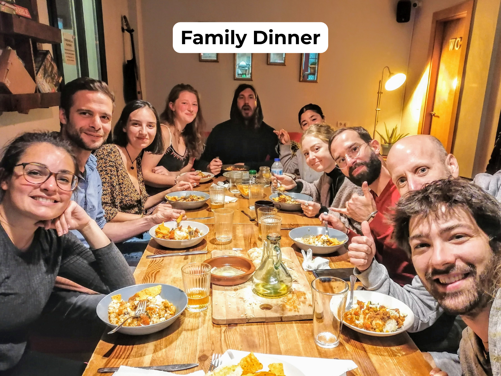 Friends gathered around a long table for a family dinner, smiling and eating in a warm restaurant setting.