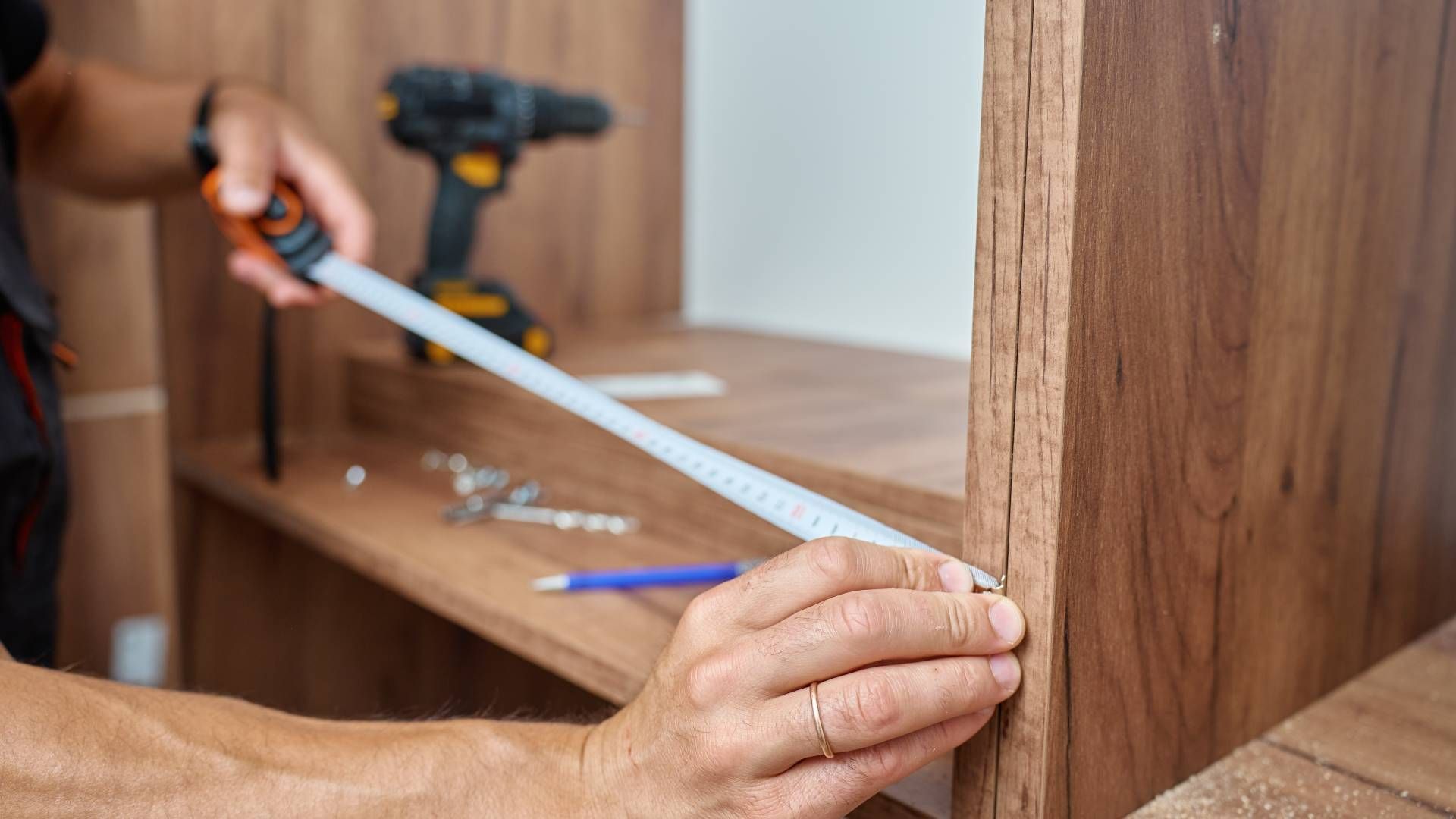 Person measuring wooden cabinet with a tape measure; drill and tools in the background.