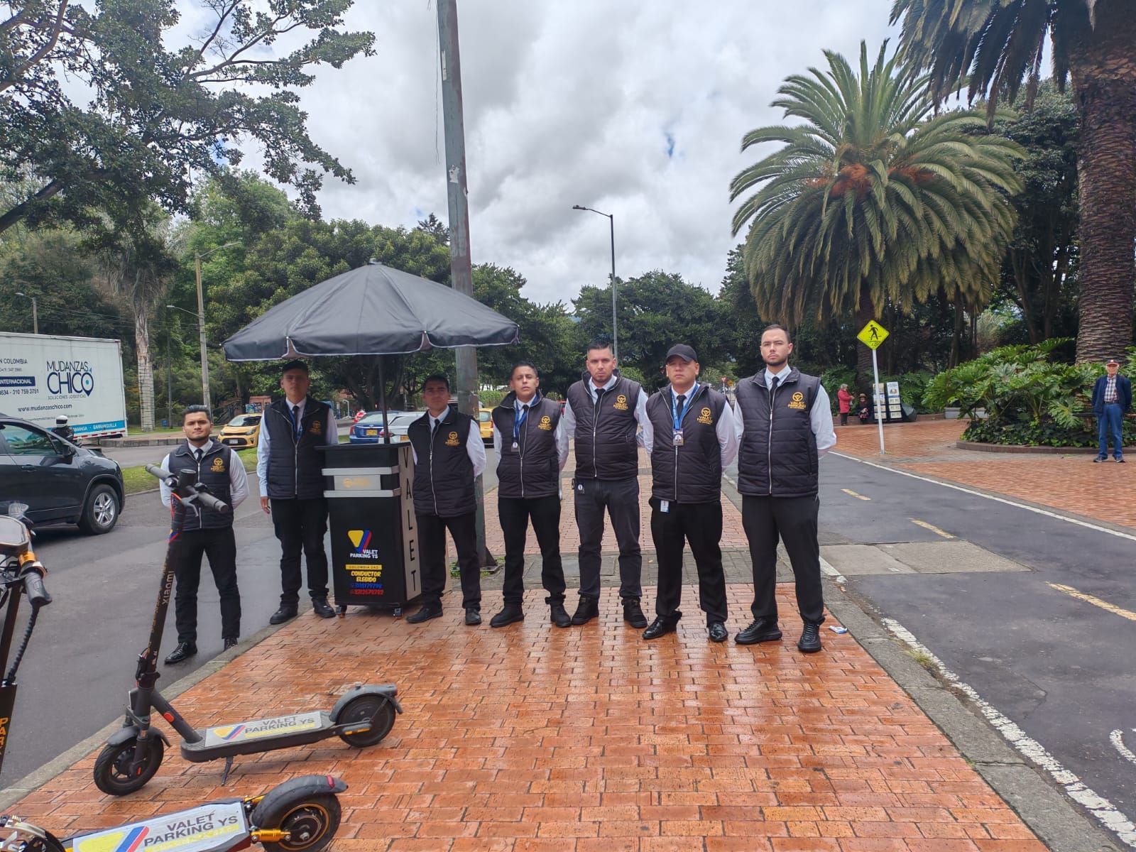Un grupo de hombres con chalecos están con scooters en una acera, bajo un cielo gris, cerca de una calle y palmeras.