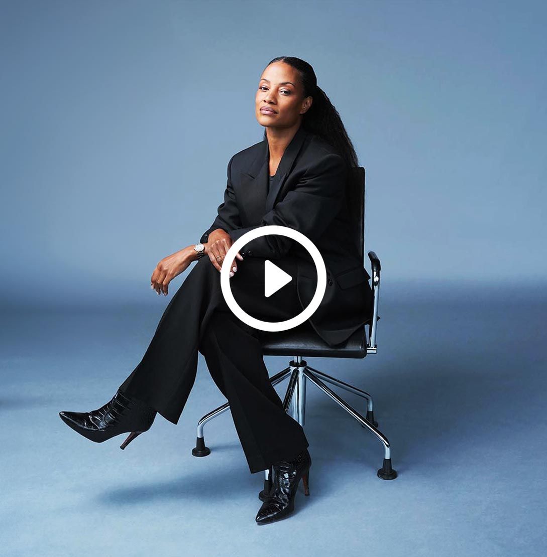 A woman in a black suit sits on a chair against a blue background. She has a relaxed pose, smiling.
