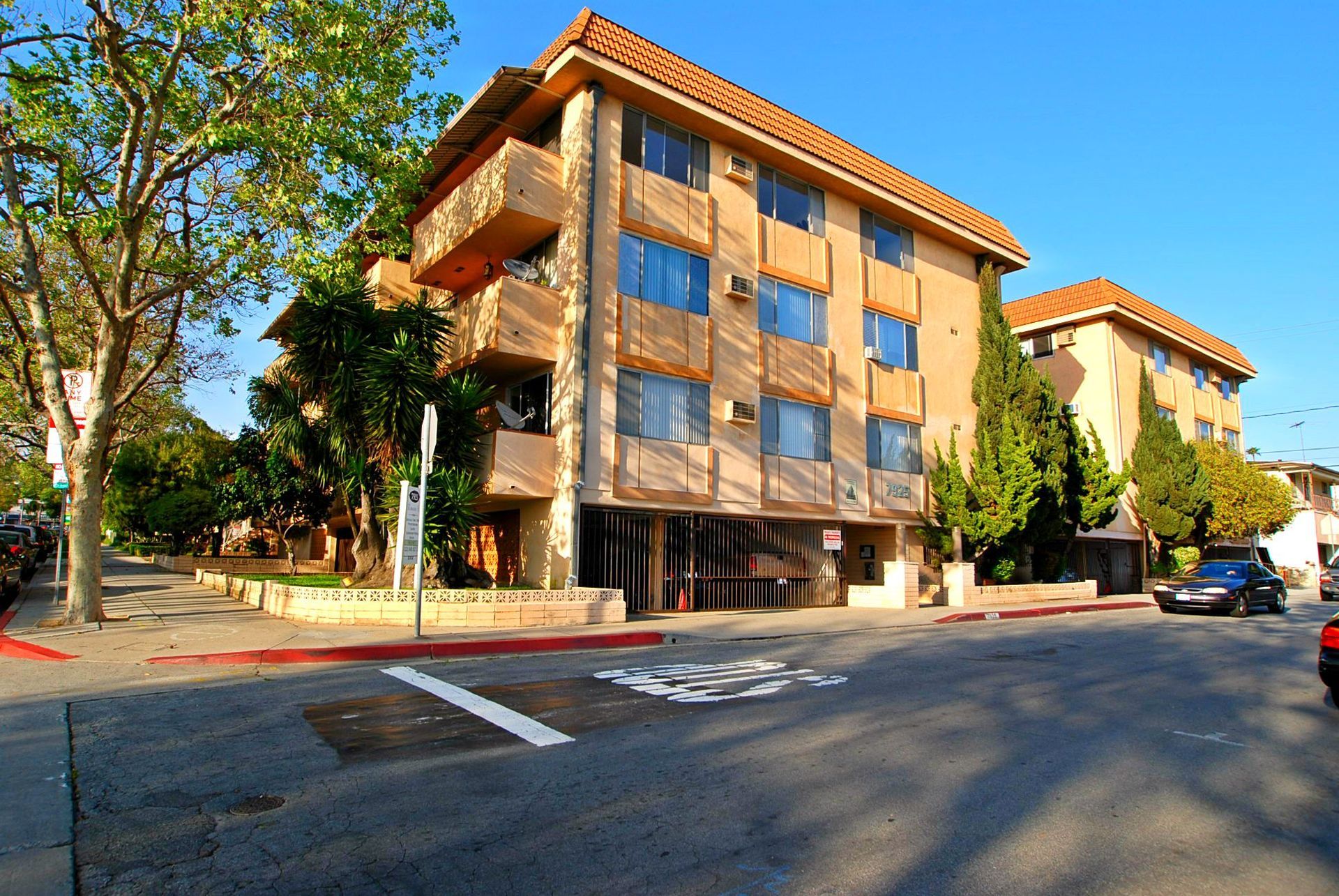 Apartment building on a street corner, tan stucco facade, tile roof, with parked cars and trees.