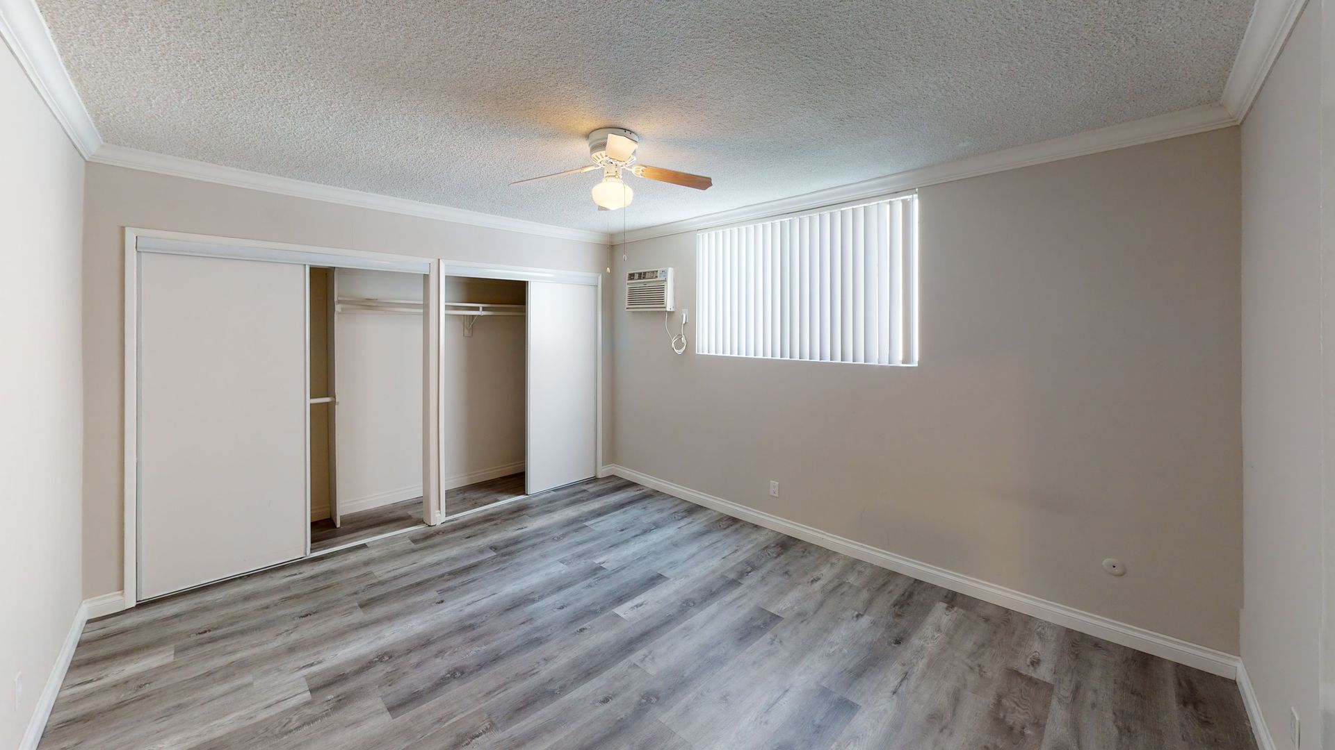 Empty bedroom with closet, window, and wood-look flooring. Light walls, ceiling fan.