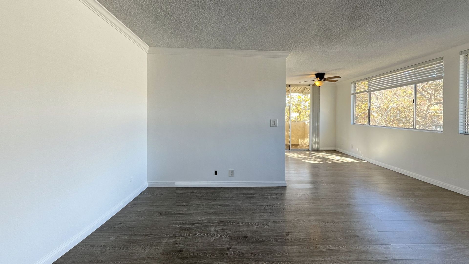 Empty, sunlit room with white walls, ceiling, and windows. Balcony visible through doorway, dark wood floors.