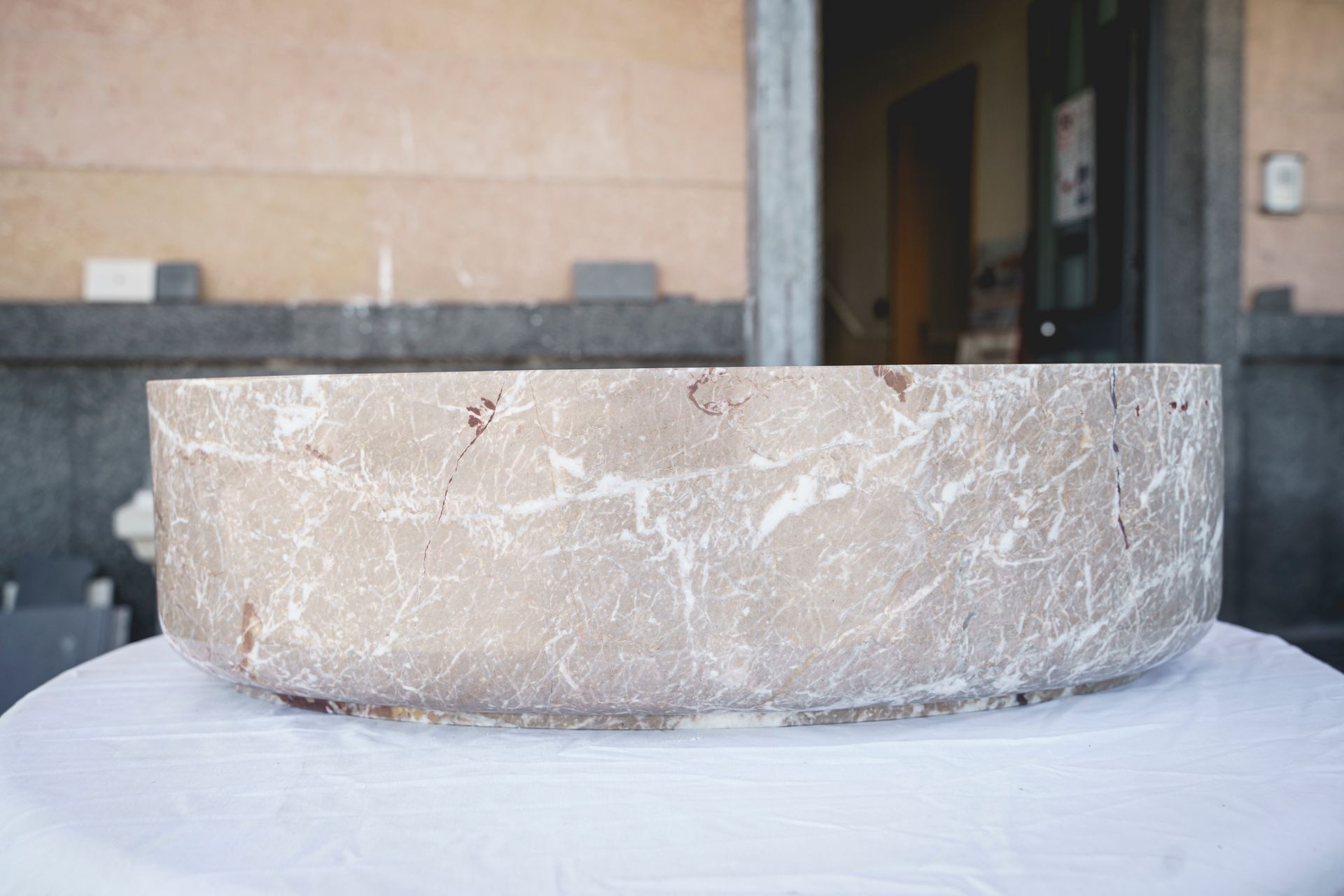 Lavandino circolare in marmo chiaro su una superficie bianca, posizionato all'aperto di fronte a un edificio.