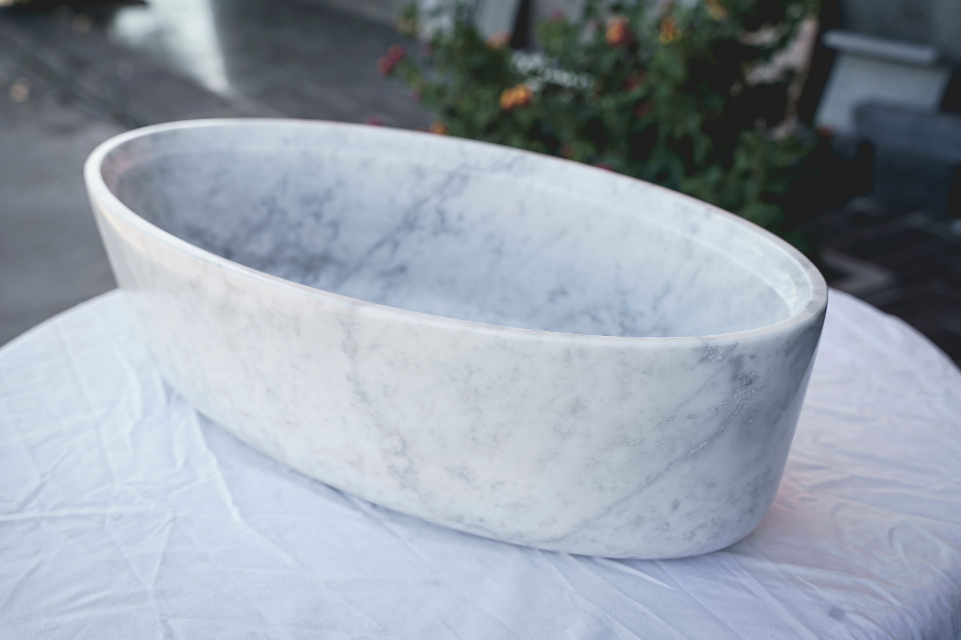 Vasca da bagno ovale in marmo su un telo bianco, posizionata all'esterno con piante sullo sfondo sfocato.