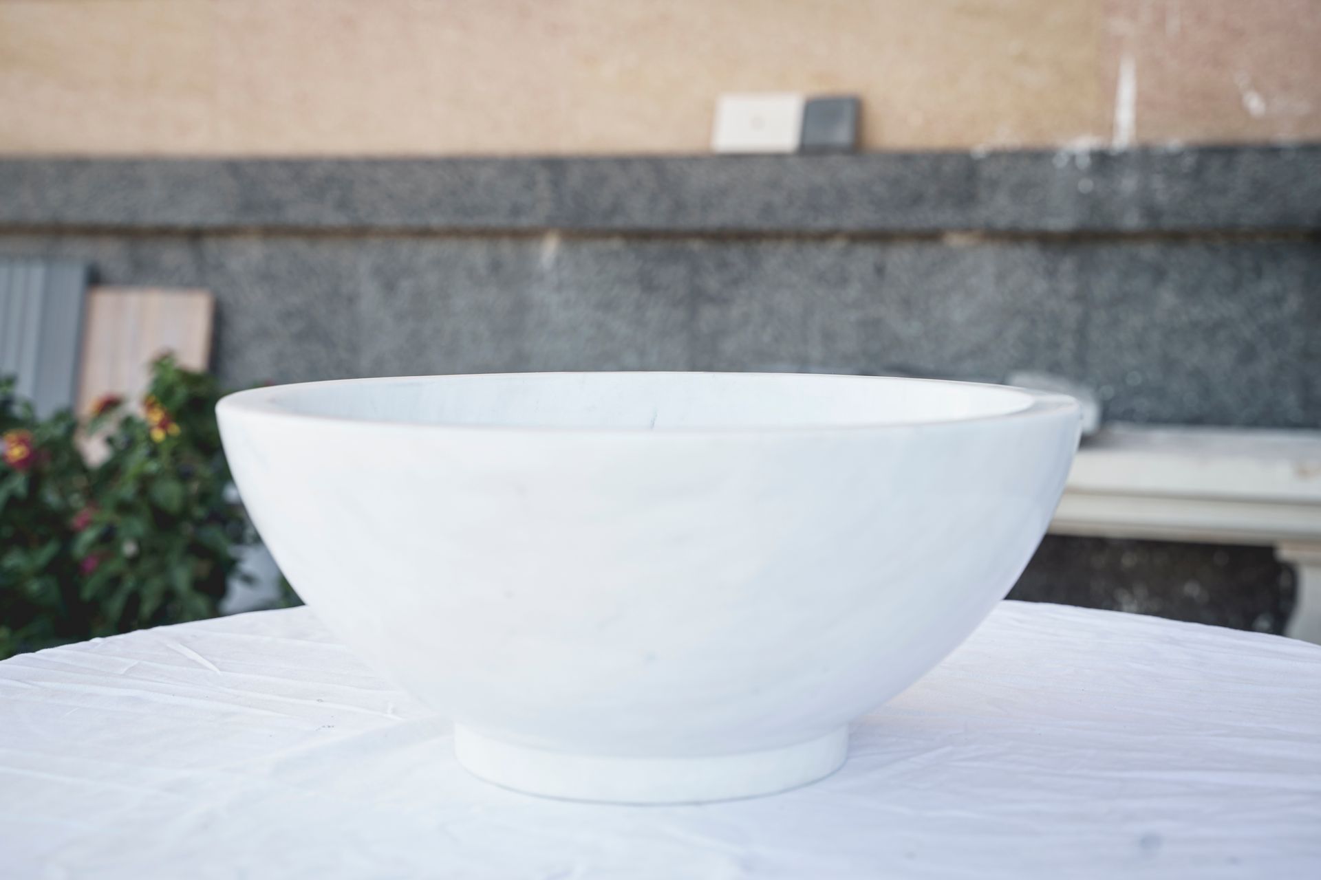 Ciotola in ceramica bianca su una superficie bianca, posta all'aperto, con sfondo sfocato di un muro e del fogliame.