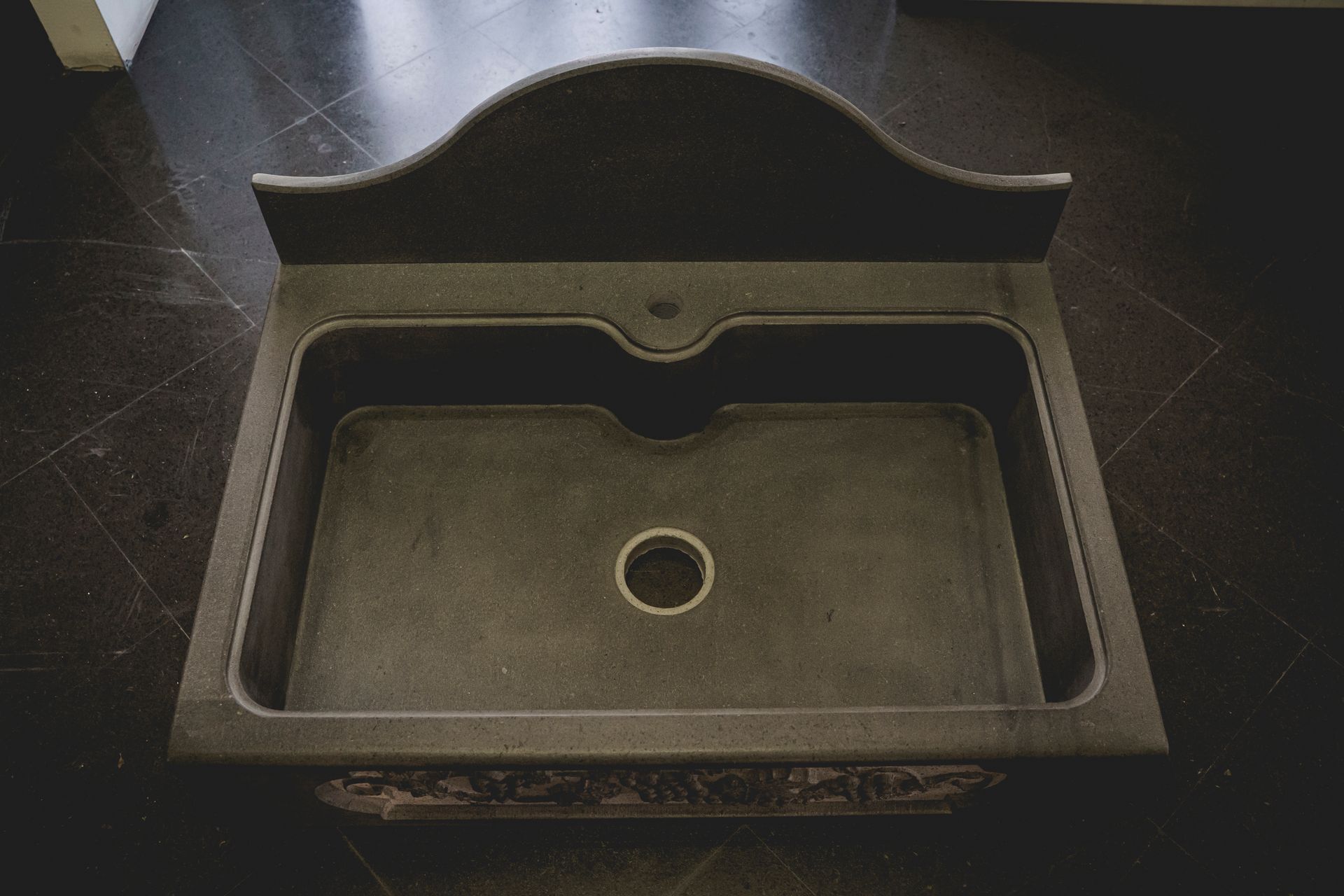 Lavello rettangolare grigio scuro in stile rustico con paraschizzi curvo, posizionato al centro del pavimento scuro.