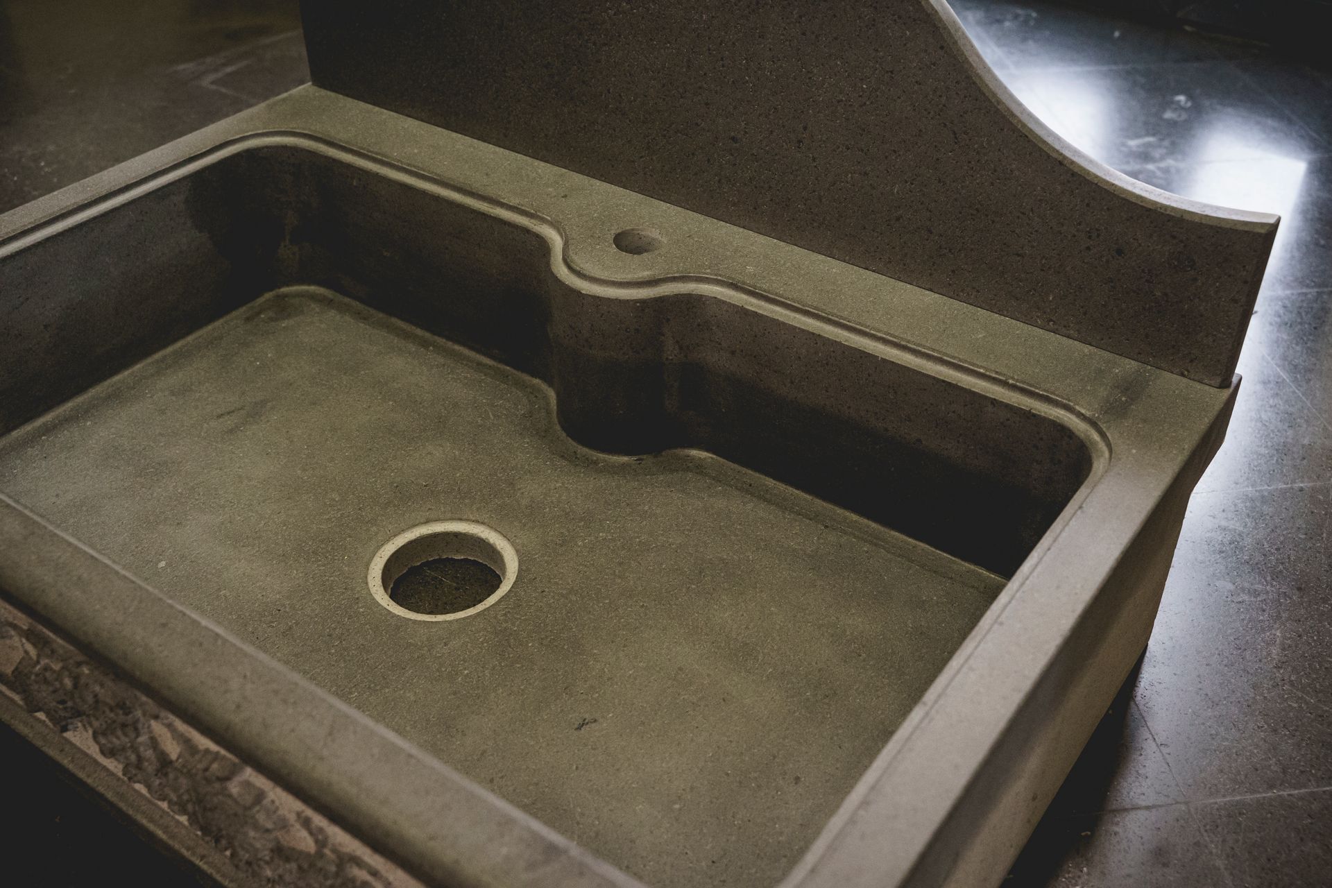 Un lavandino in cemento grigio con paraschizzi curvo e foro per il rubinetto, inserito in un ambiente buio.