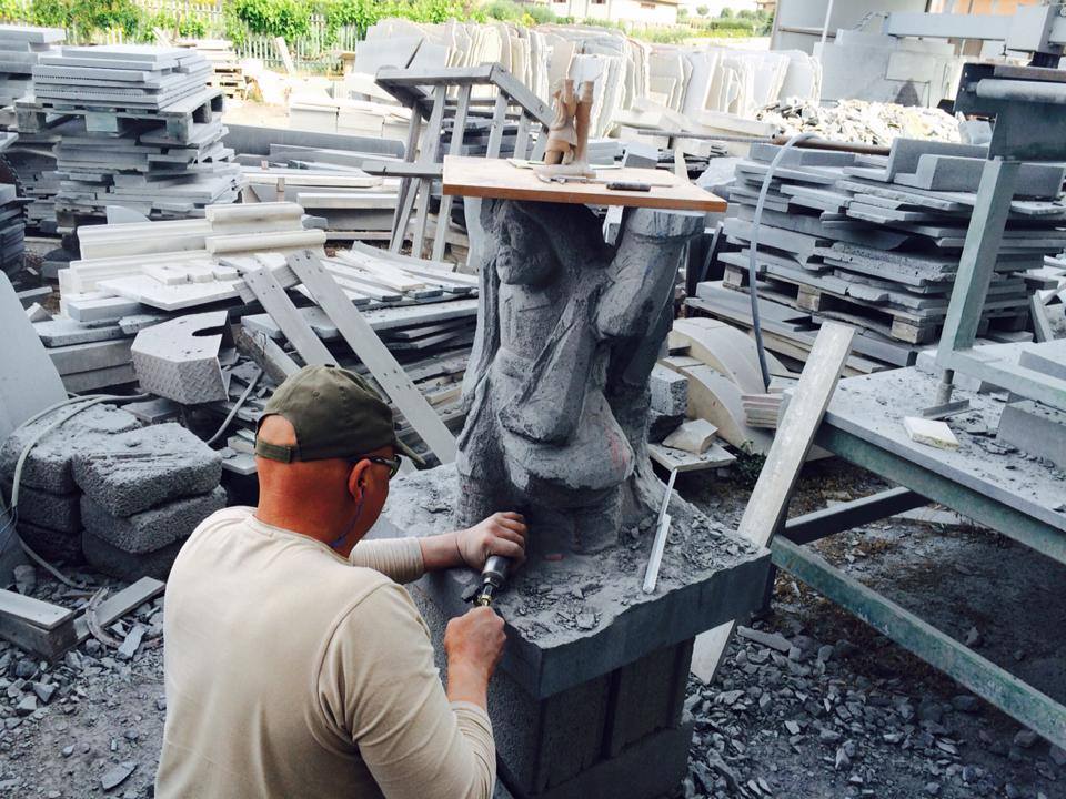 Scultore che scolpisce una statua in pietra all'aperto, con gli attrezzi. Ambiente di laboratorio con pile di pietre.
