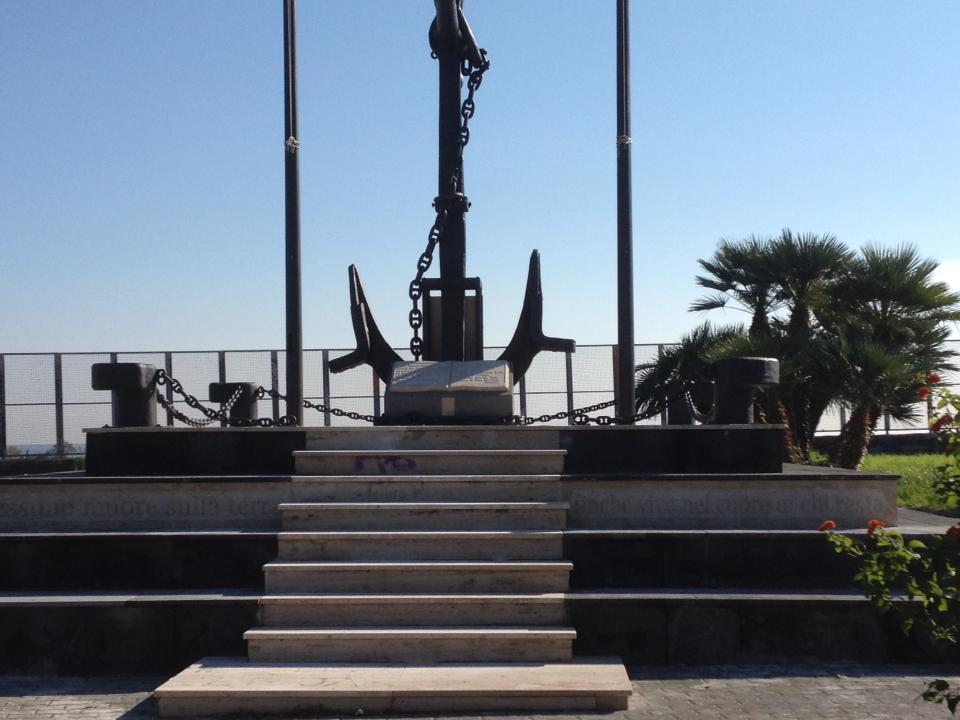 Monumento con grande ancora, catene e gradini, in una giornata di sole con palme sullo sfondo.